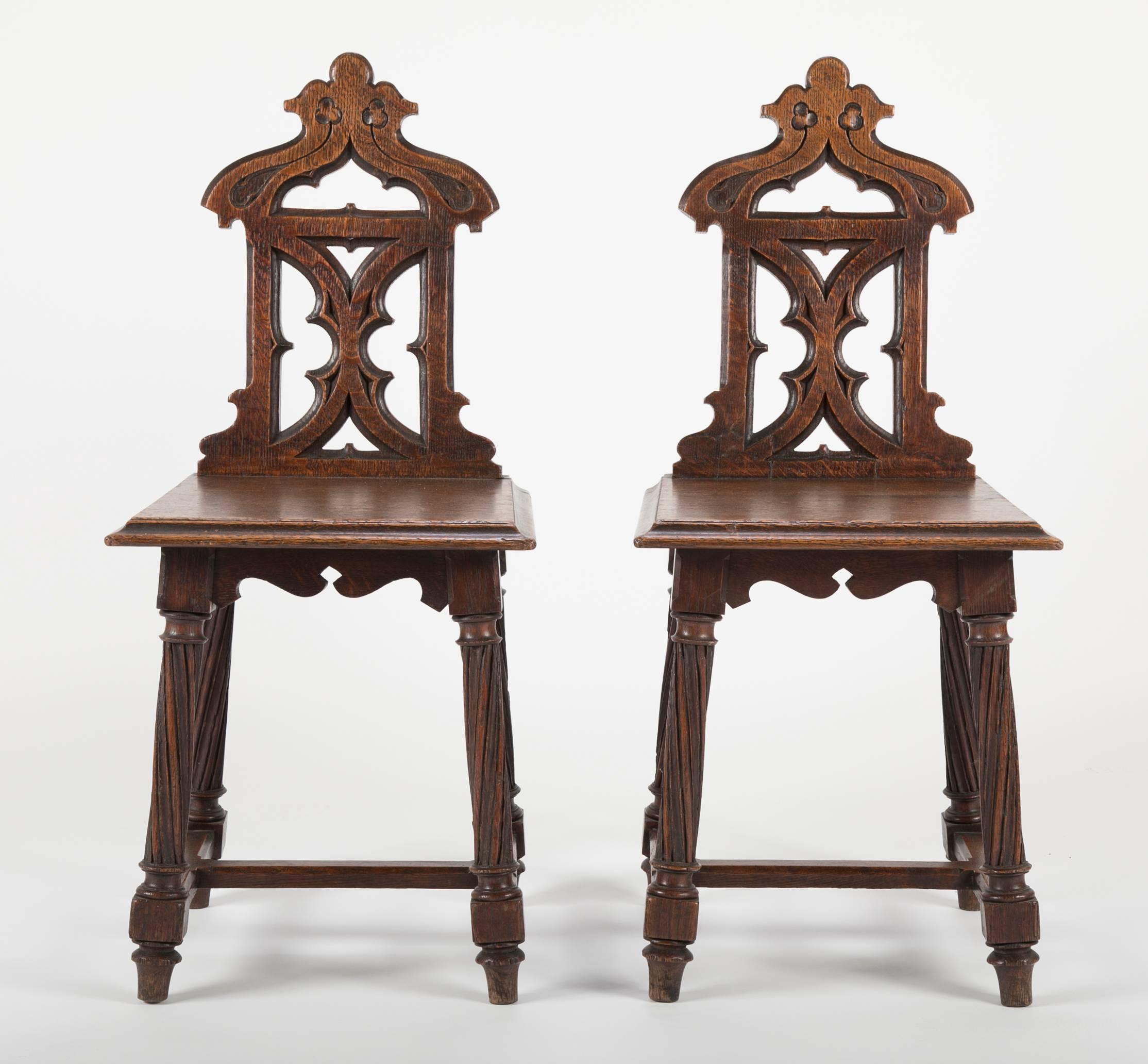 A fine pair of English oak hall chairs with Gothic tracery back splats topped by incised trefoil decoration, supported by four turned legs joined by stretchers with chamfered edges.