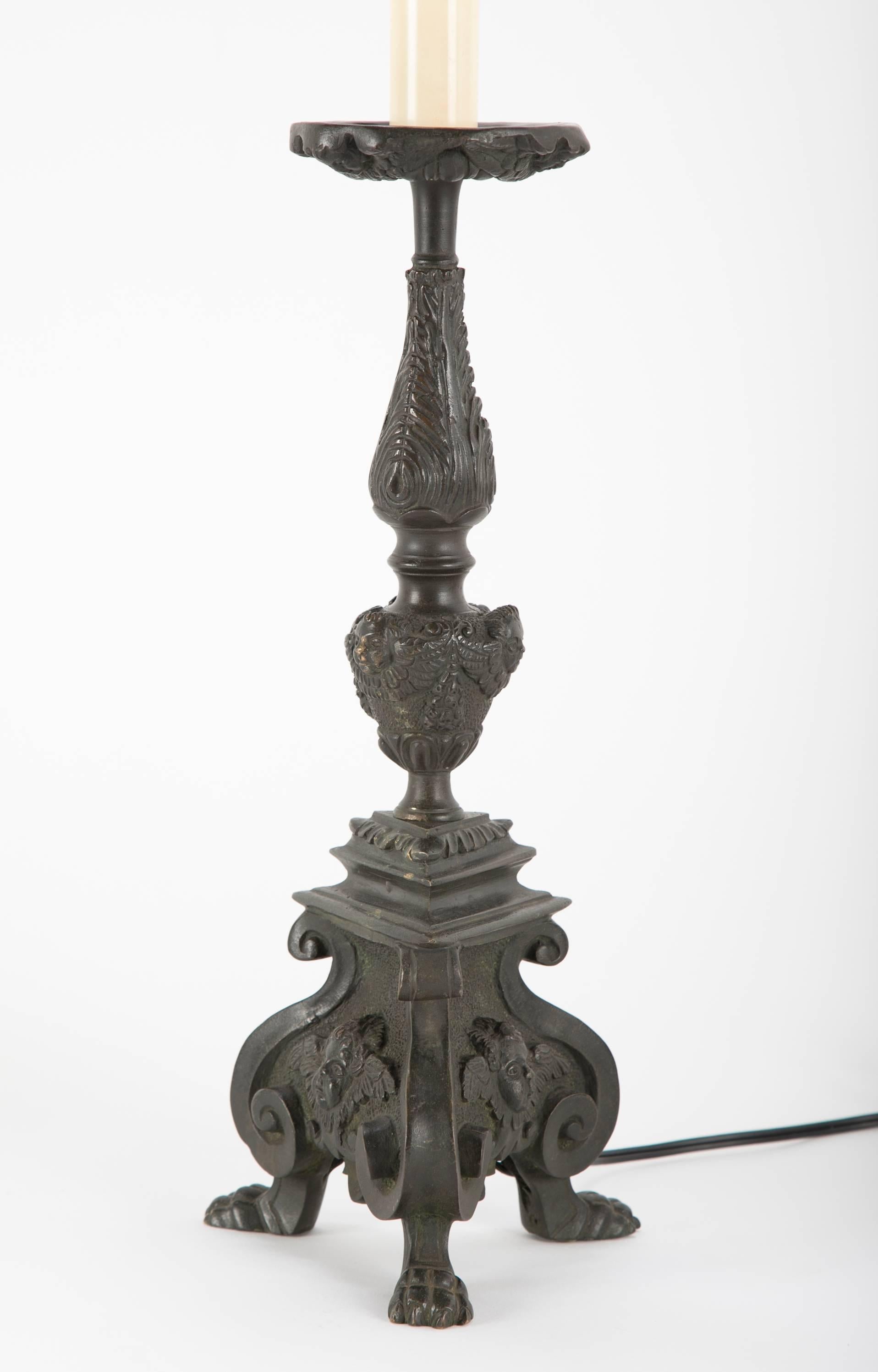 Pair of Italian Baroque style bronze candlesticks wired as table lamps. With tripod footed bases decorated with winged putto faces on each side. Additional putti on the center knob and the underside of the drip pan. Deep greenish back patina. With