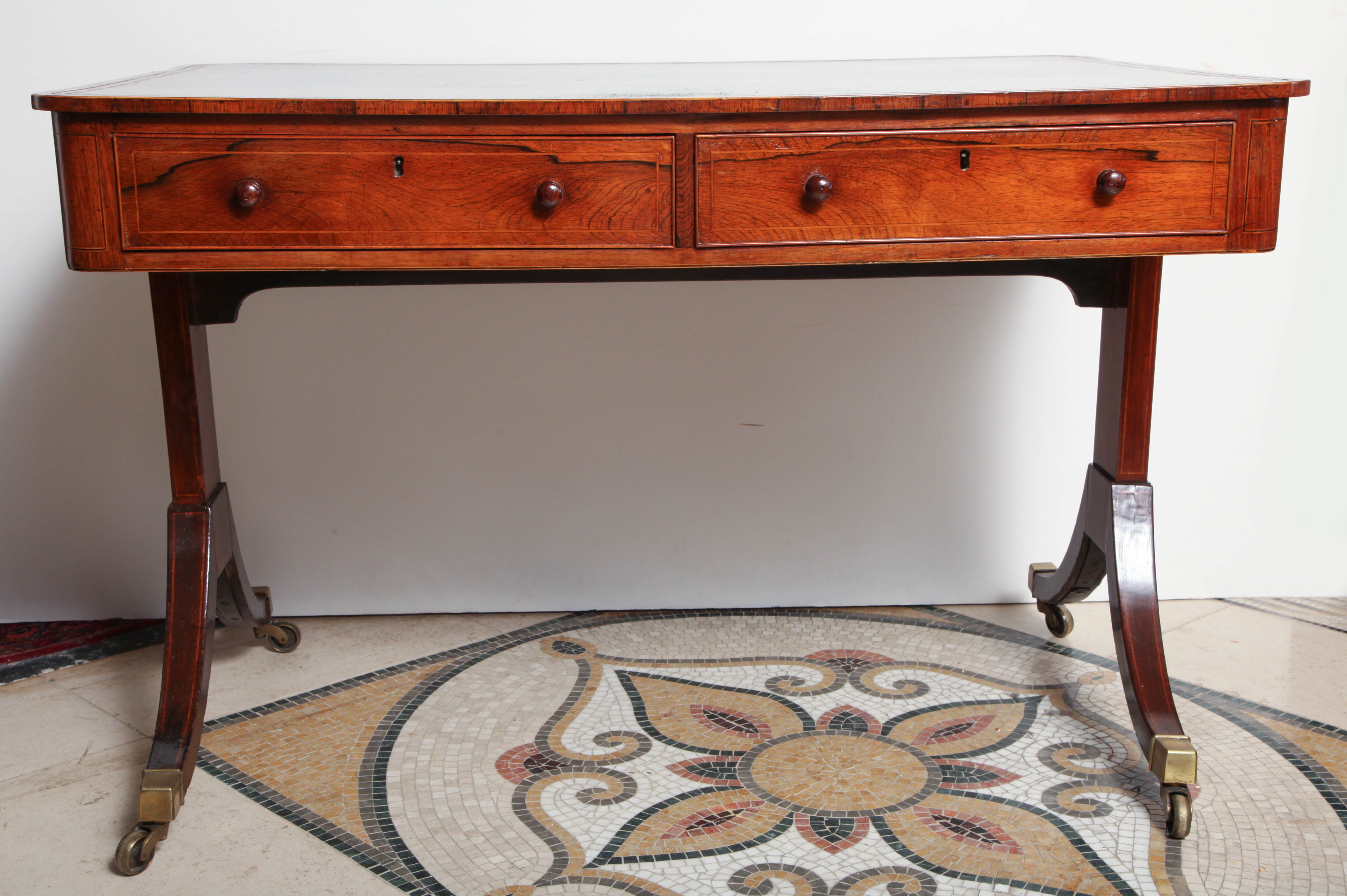 A Regency rosewood trestle-end writing table with boxwood stringing and an inset leather top above a two-drawer frieze, the splayed legs ending in brass casters.