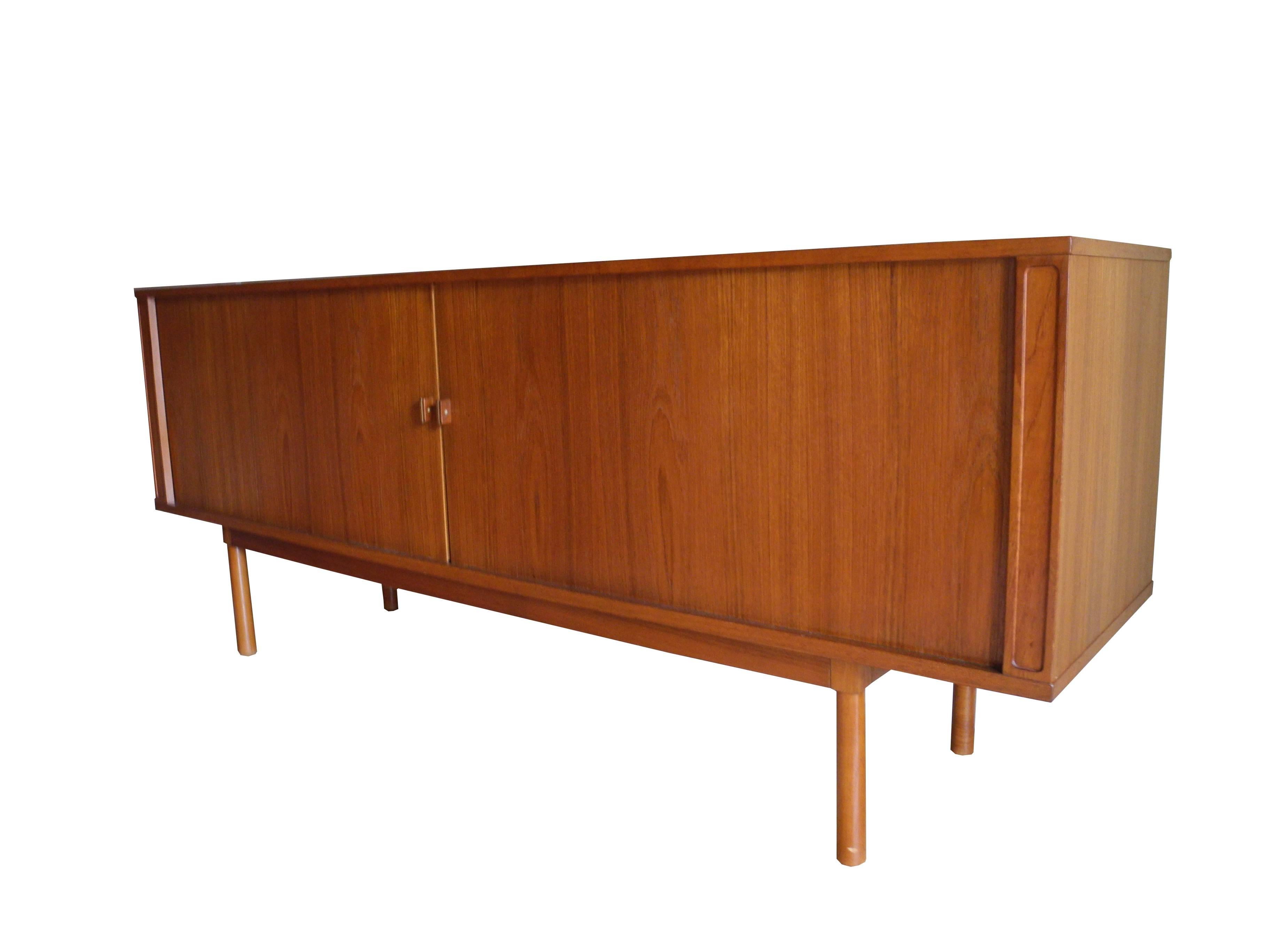 Stamped Løving, this Danish Modern, tamboured door sideboard, presents with a large amount of storage space. The side shelves are adjustable. The teak has a warm glow and the knobs have a brass accent.