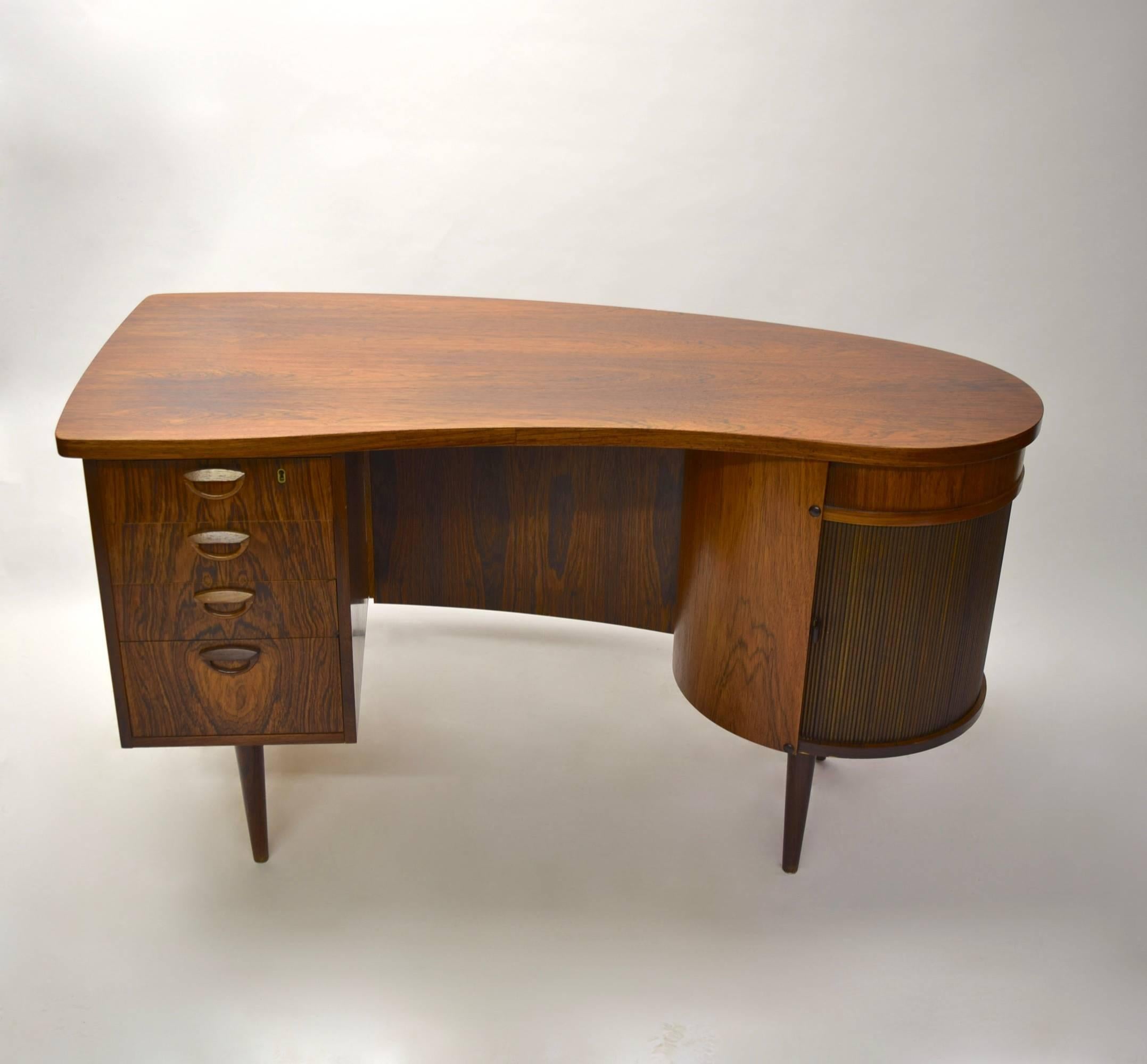 Desk in rosewood has four drawers on the left side each with matching wood pulls. On the round right side is a home bar behind a tambour door, above is a concealed drawer. 
Model 54. Designed 1956 by Kai Kristiansen for FM Furniture, Feldballe