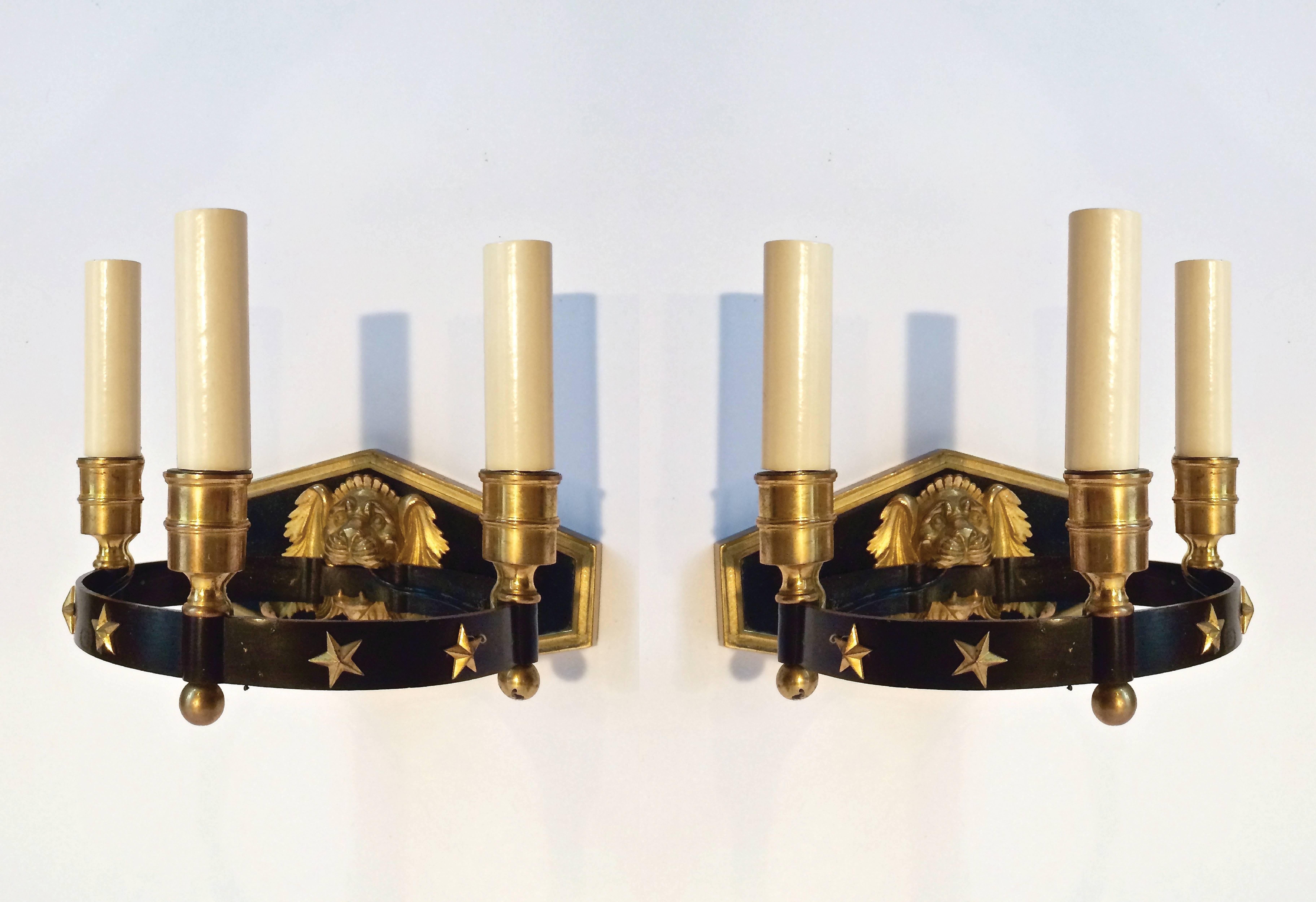 Une paire d'appliques classiques à trois lumières, inspirées d'anciennes appliques Empire. Chacune d'entre elles comporte une armature circulaire contenant trois lumières et décorée d'étoiles en laiton, reliée à un support en forme de visage de lion