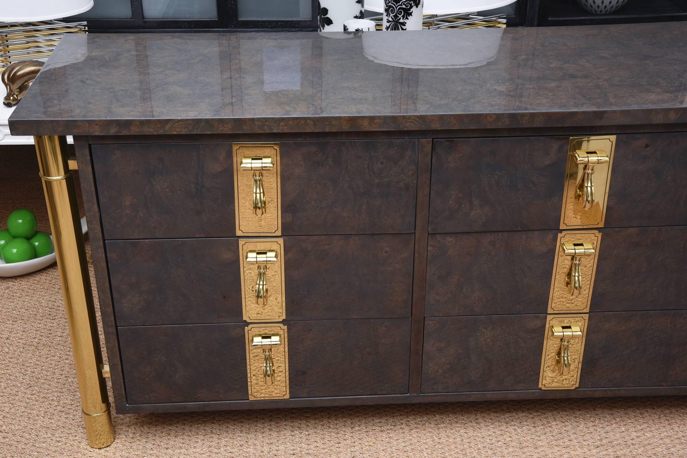 This fabulous newly restored Mastercraft burl elm wood dresser has heavy substantial solid brass knockers as the pulls and the side panels and column legs are also heavy brass. They have all been polished to beauty. There are nine drawers. The