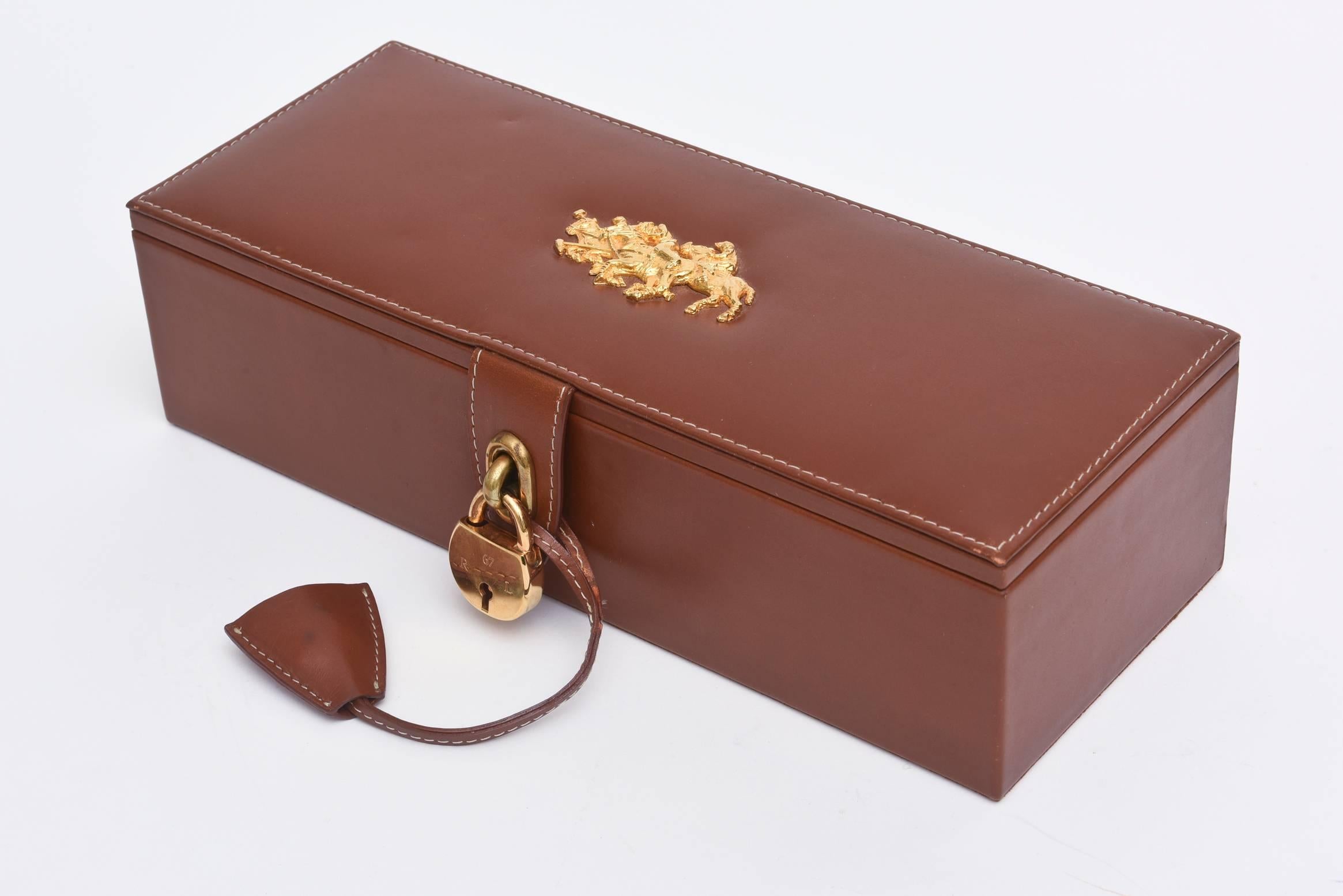 Ce superbe coffret en cuir souple marron surpiqué de Ralph Lauren est doté d'une quincaillerie plaquée or représentant le motif d'un match de polo. Il est doté d'une serrure et de deux clés logées à l'intérieur de la pochette en cuir. Il abrite cinq