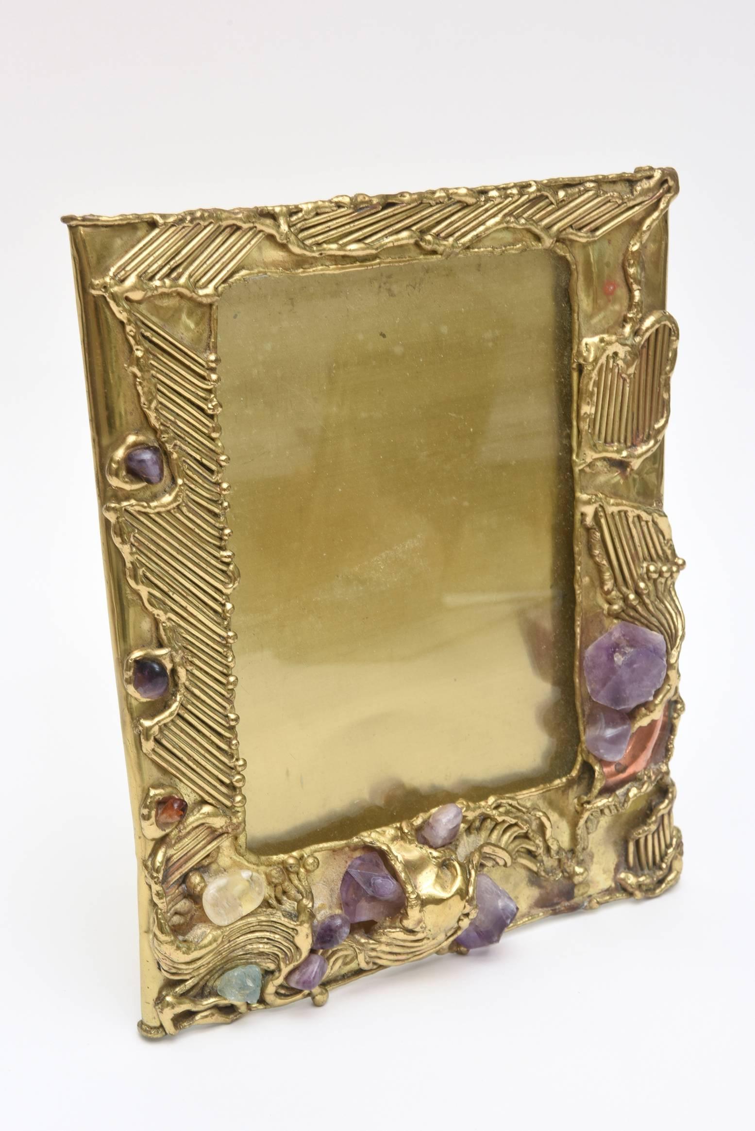 This studio one of a kind hand-wrought textural and dimensional picture frame is a mixed media of brass with inlays of copper hints.
The jeweled like frame/mirror has dimensional juttings of raw amethyst, rose quartz and agate. It is sculptural and