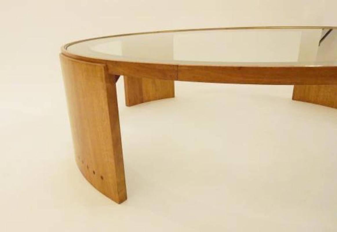 Large scaled round Mid-Century cocktail or coffee table featuring a body in oak with four large curving leg supports each inset with a line of dark wood plugs at the foot. The table also features an inset glass top edged with a gilt brass band. The