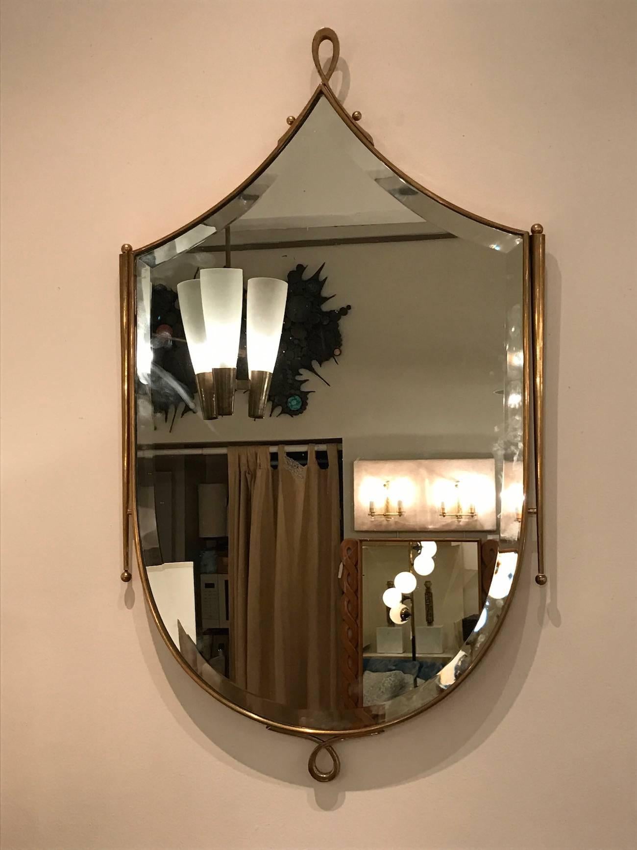 Mid-Century wall mirror with a clear mirror with a wide bevel featuring a shield shaped body framed in brass with an upper and bottom loop design. The frame also features tapering side batons with ball finials. In the style of Rene Prou, France,