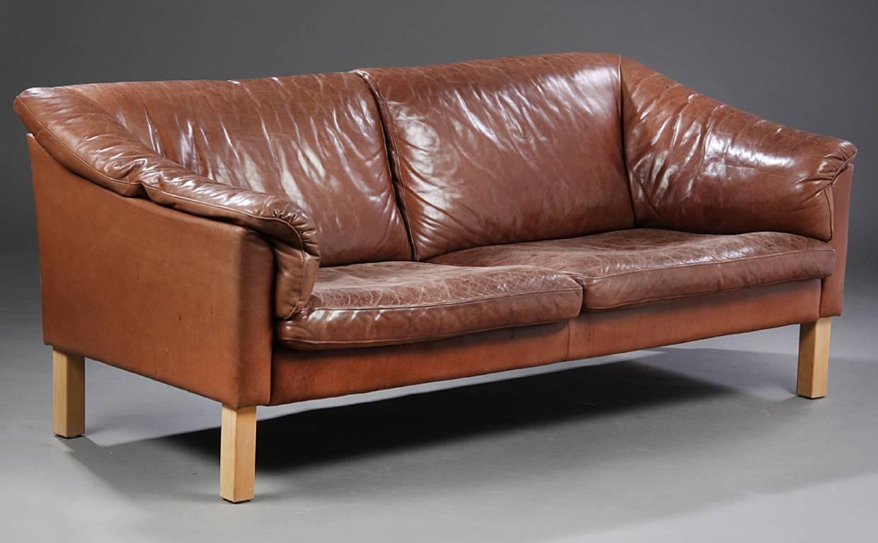 Pair of Danish 1960s-1970s loveseats upholstered in a very good quality leather. The leather of a beautifully aged tobacco color. The loveseats raised on beechwood legs. Leather is in very good presentable antique condition, but leather is vintage