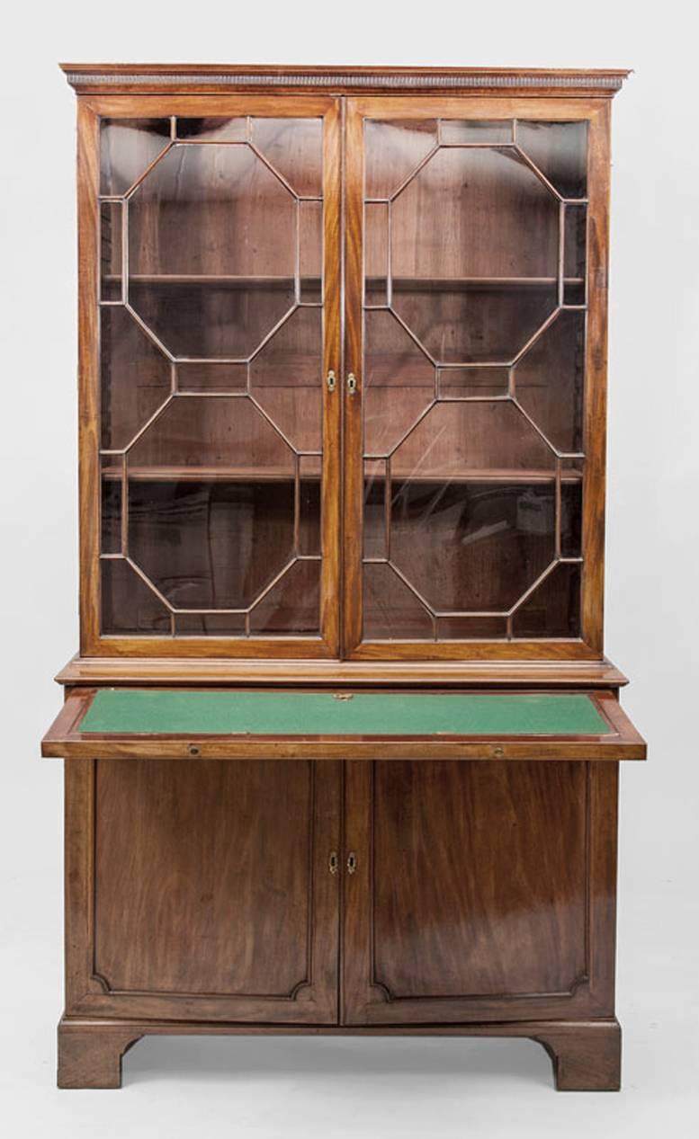 George II two-part mahogany bookcase. Mid-18th century. The dentil molded cornice over two glazed doors opening to an interior fitted with shelves. The doors over a pull out fall front with a baize lined ratched writing surface that slants up to