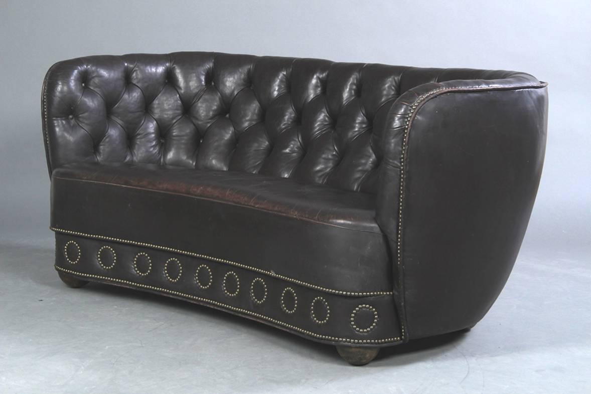 1930s-1940s Danish banana form loveseat upholstered in a Chesterfield style with a bottom frieze mounted with circular close-nailed decoration. The loveseat raised on ball feet. A good apartment sized sofa.