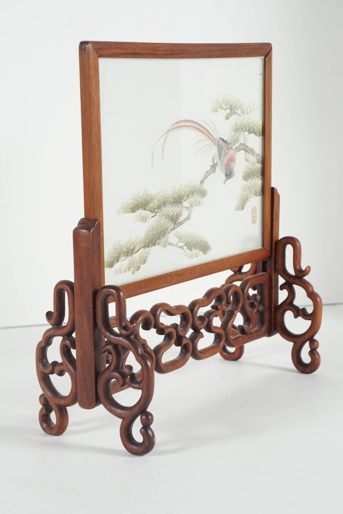 This early 20th century screen is refined and very beautiful. The image of a bird observing life from a tree is worked in thread on silk mesh sandwiched between two sheets of glass. The panel is signed and sits in a nice rosewood stand. The mesh has