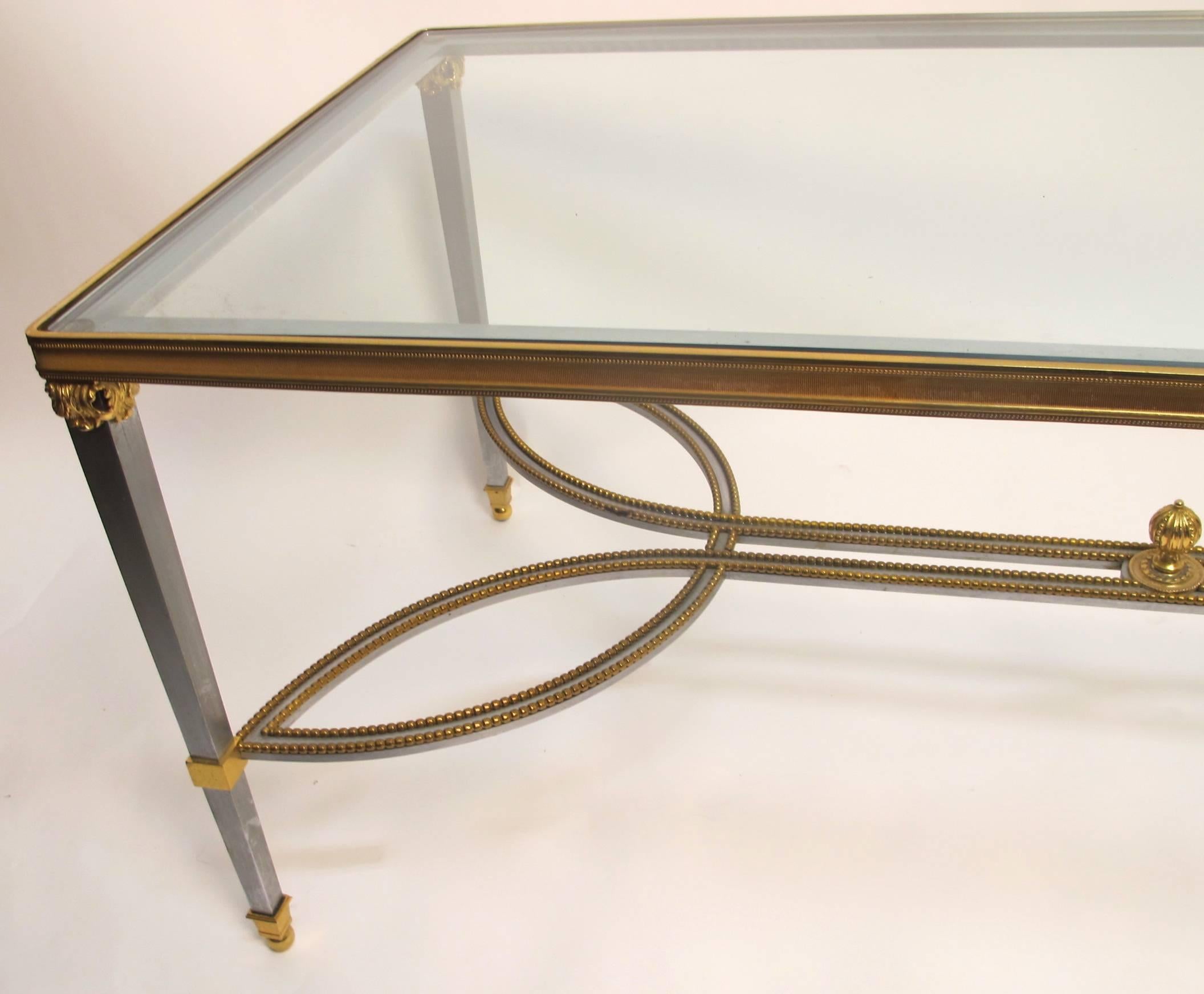 Neoclassical Revival style steel and gilt brass trim coffee table with inset glass top. This extremely well made piece has a number of interesting design features including a beaded detail and finial on its stretchers and an acanthus leaf capitol