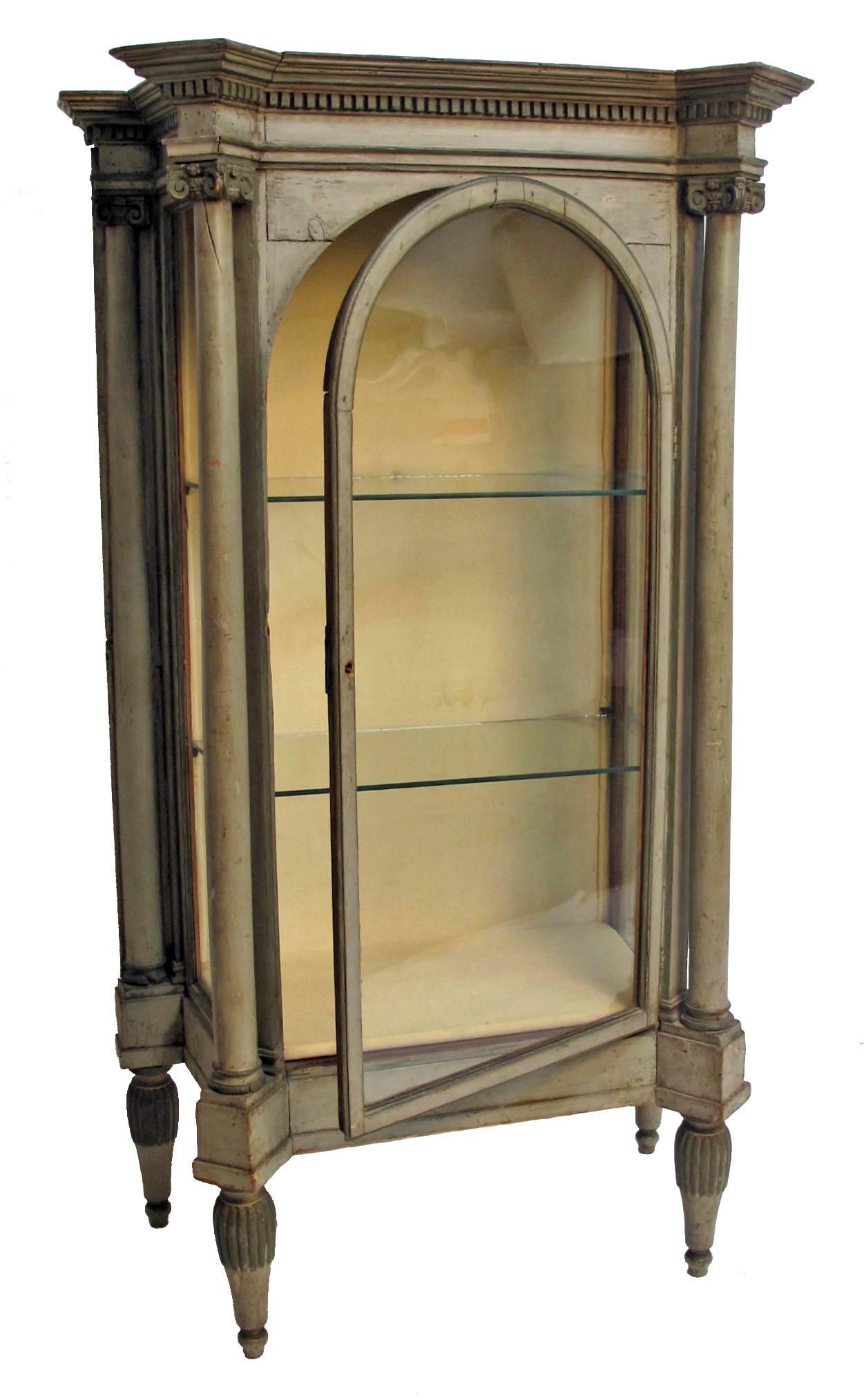 Charming green painted display case or vitrine, neoclassical with carved moulding above pair of corinthian capital columns flanking the arched glass door and pair of pilasters flanking the angled side windows. Standing on tapering cylindrical legs.