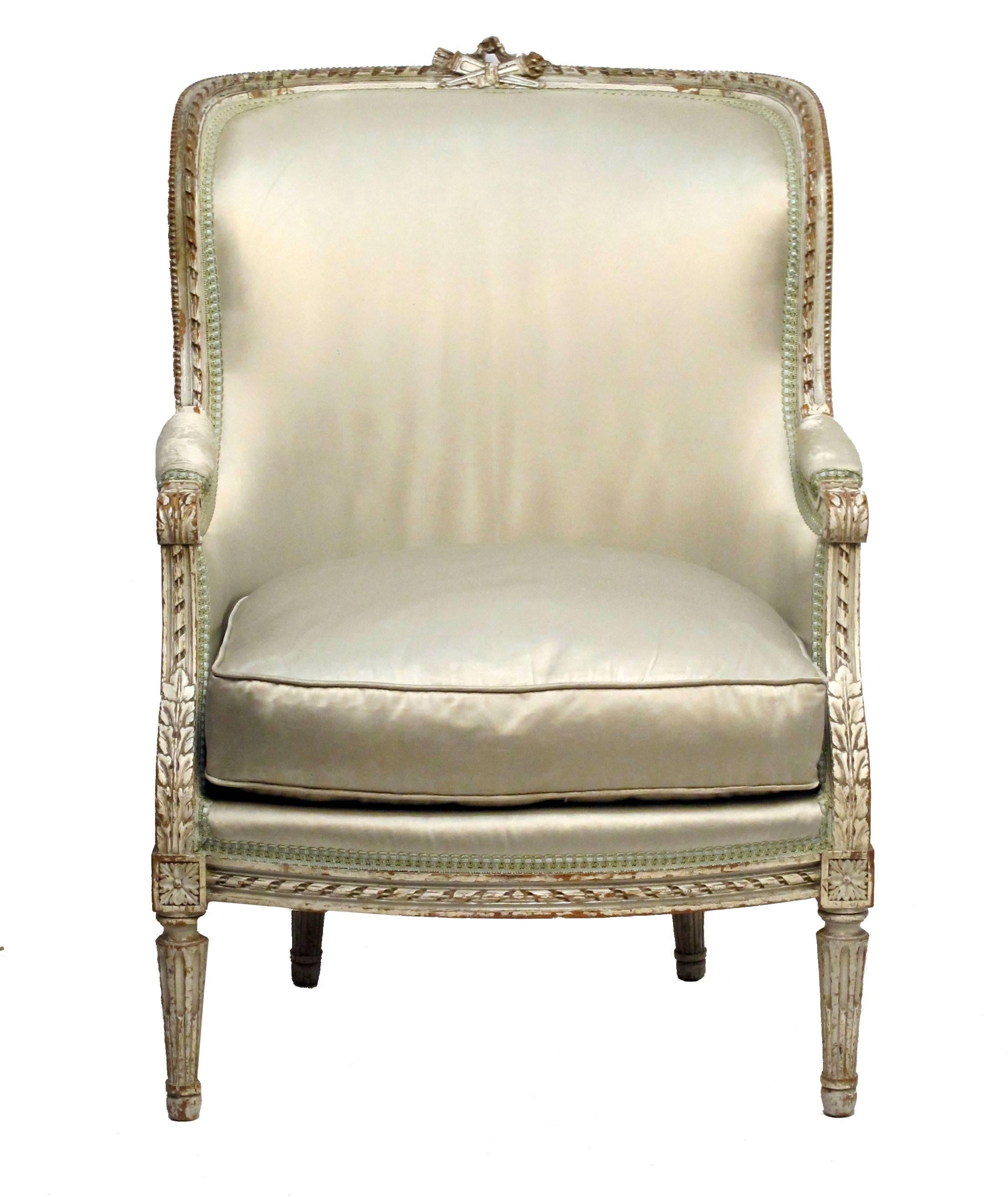 Elaborately carved and painted Louis XVI style bergere chair. The light green painted frame carved with twisted rope and curved back surmounted with crossed wheat and quiver design, standing on tapering fluted legs. Older satin upholstery in good