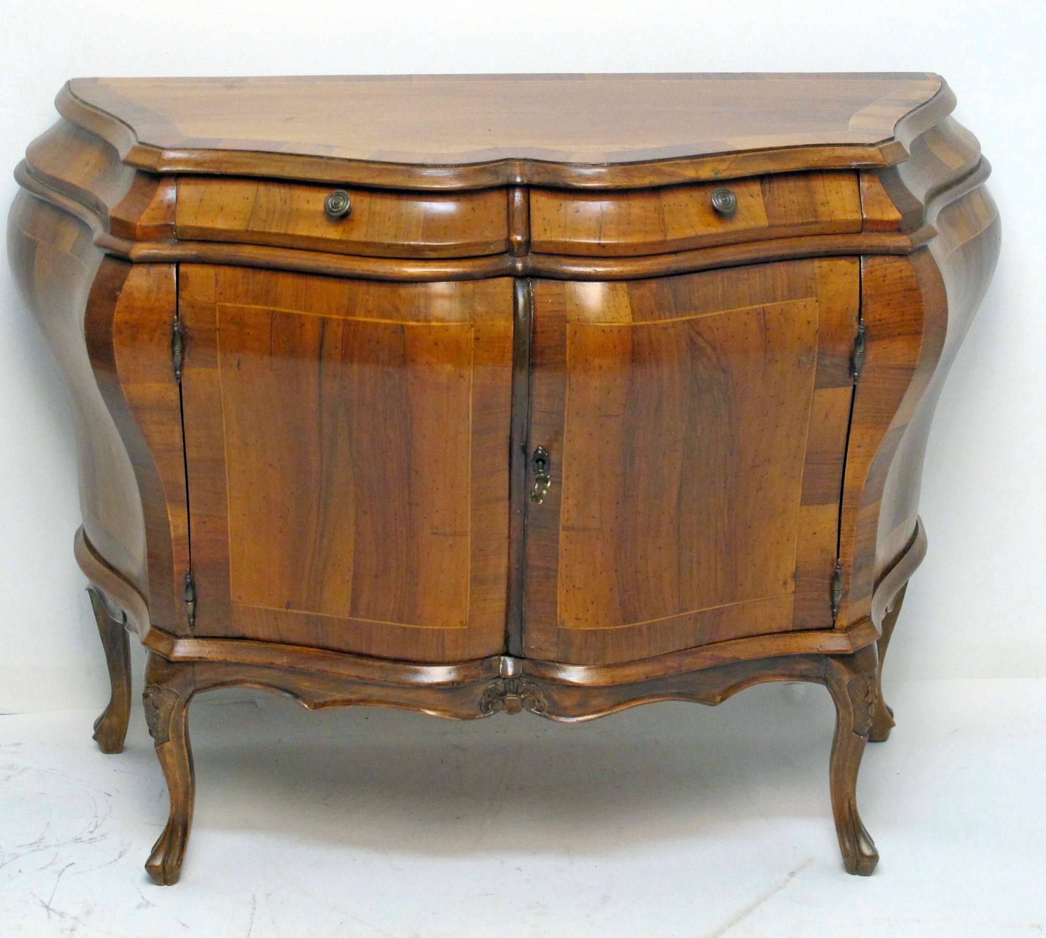 A robust vintage walnut bombe chest with mahogany crossbanding and satinwood inlay. This commode has two short drawers above pair of cabinet doors, opening to interior shelf standing on cabriole legs with decorative carving to the knees; with key