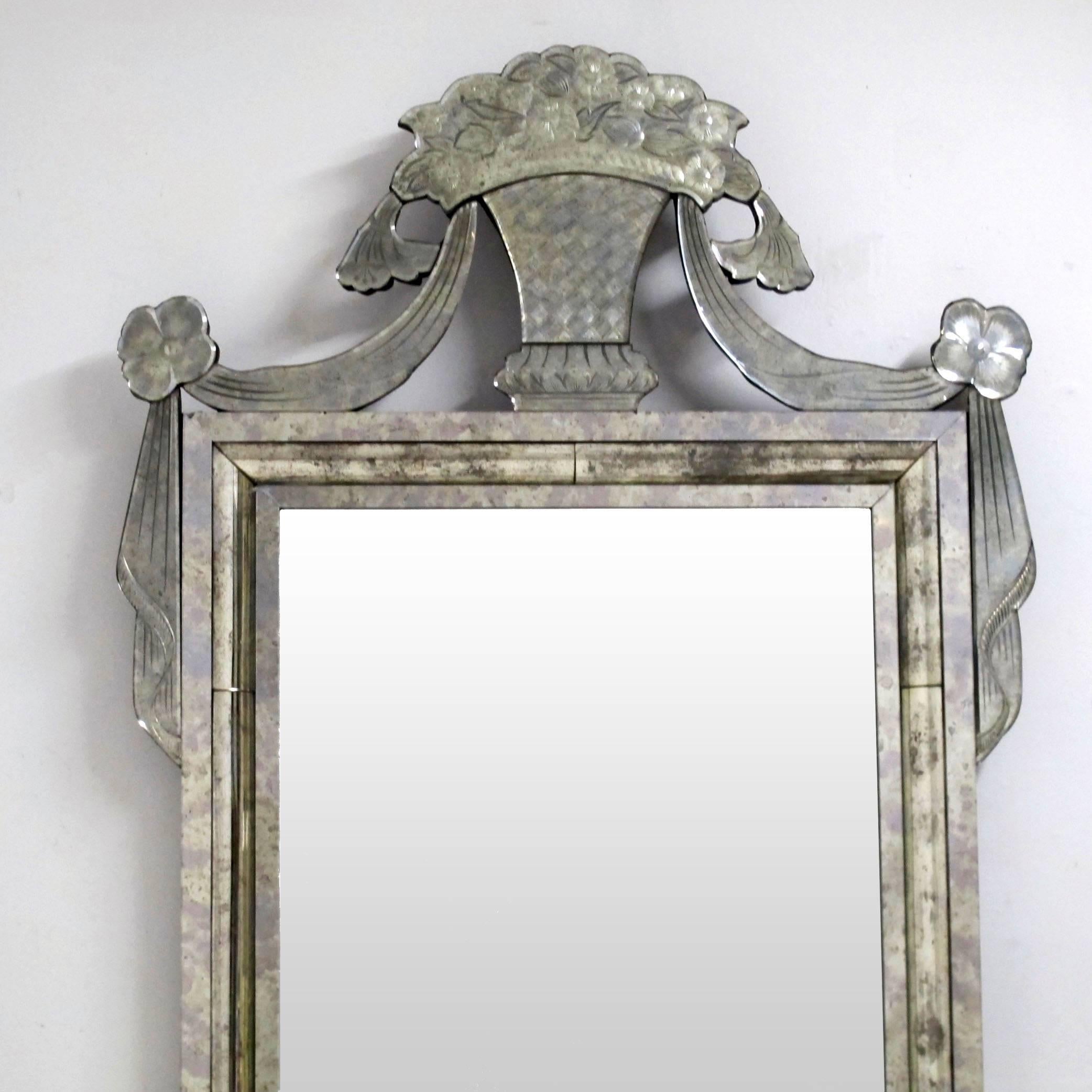 A flamboyant Venetian style mirror with beveled and aged glass frame, having an etched glass flower basket at the top with draped swags. American, 1920s.
