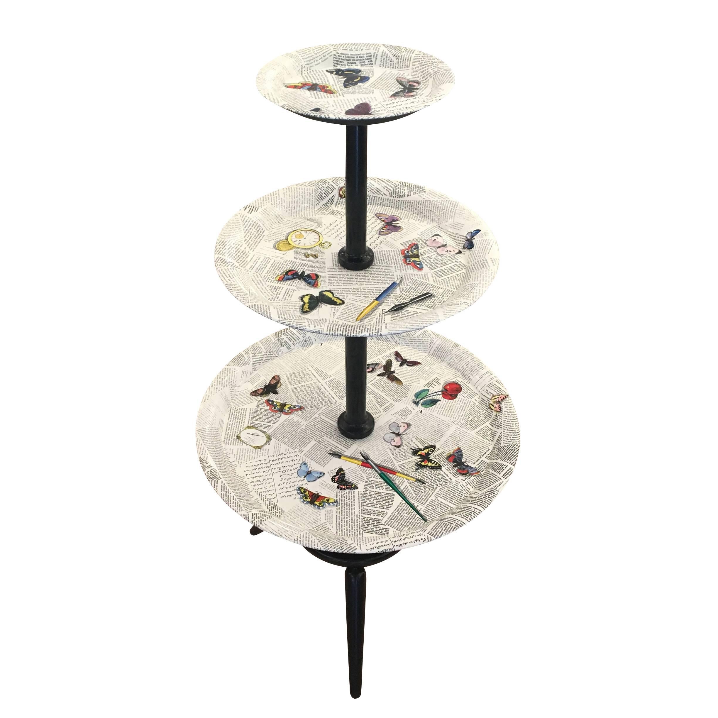 Elegant three-tiered table by Piero Fornasetti featuring a newspaper and butterfly motif. The newspaper motif is created by many overlapping articles about Fornasetti in different languages. Fun and curious to read! The three trays are metal while