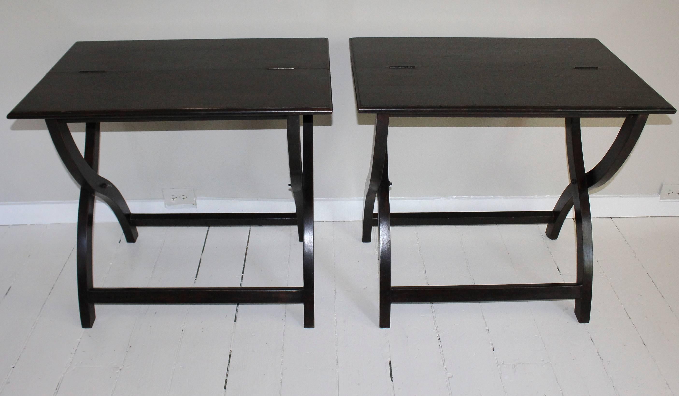 Matched pair of ebonized folding campaign-style or coaching tables, hinged at the centre to fold and stow away when not in use, supported on X-frame base with cross stretchers.