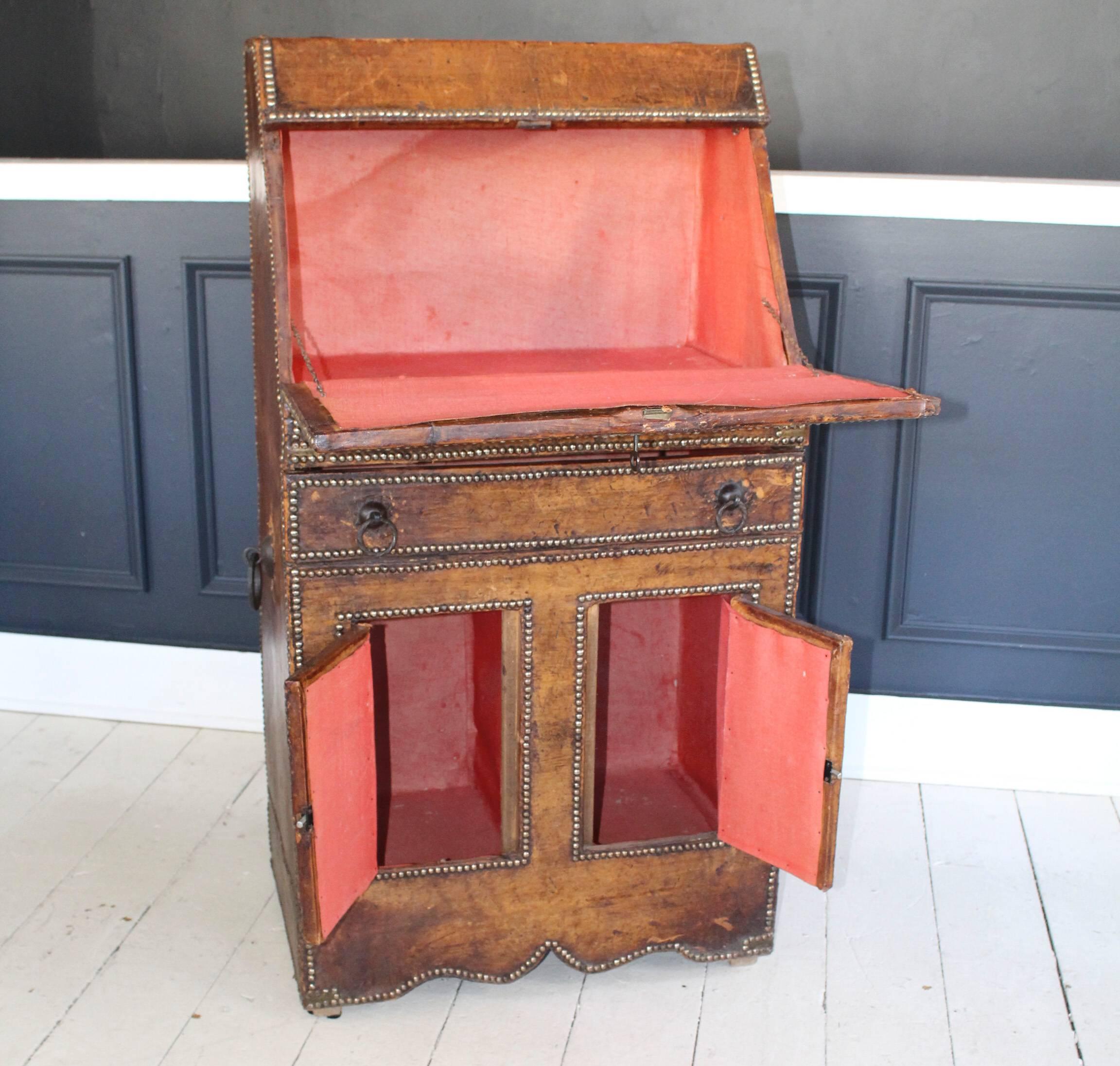 Leather slant top desk, 19th century, France, with single drawer atop two-door cabinet. Desk is outlined with nailheads. Iron ring drawer pulls and side handles add further decoration as does brass door hardware and escutcheons. Interior lined with