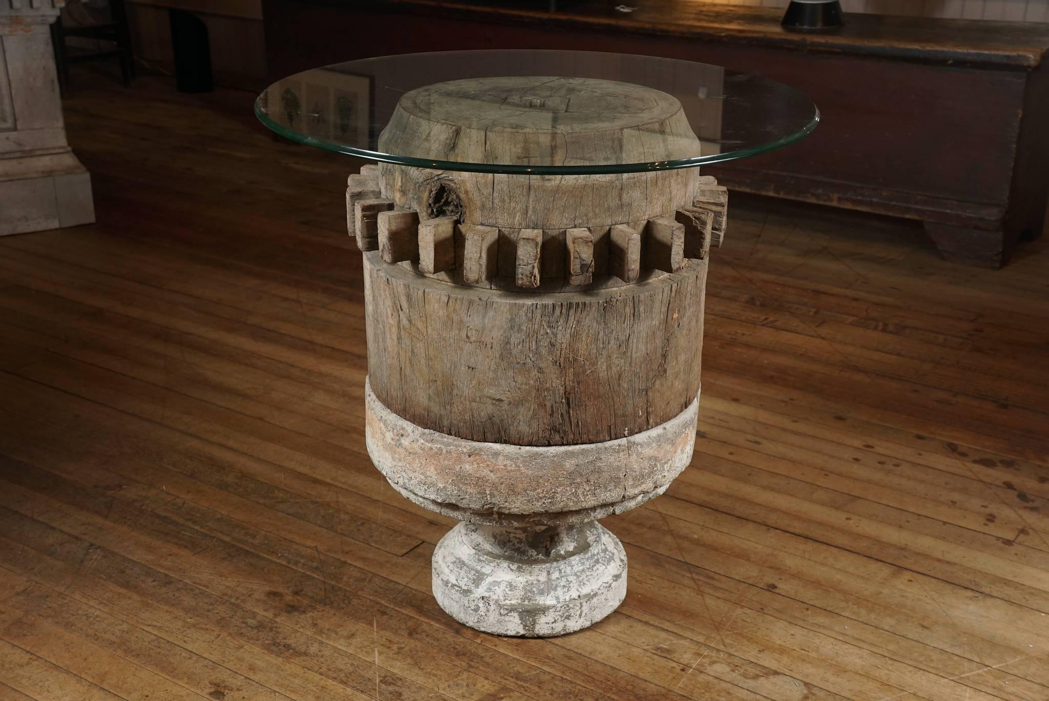 Ancient wood gear in maple, allegedly from a water mill, retains collection number and rests on a white distressed cement base to support a glass top.