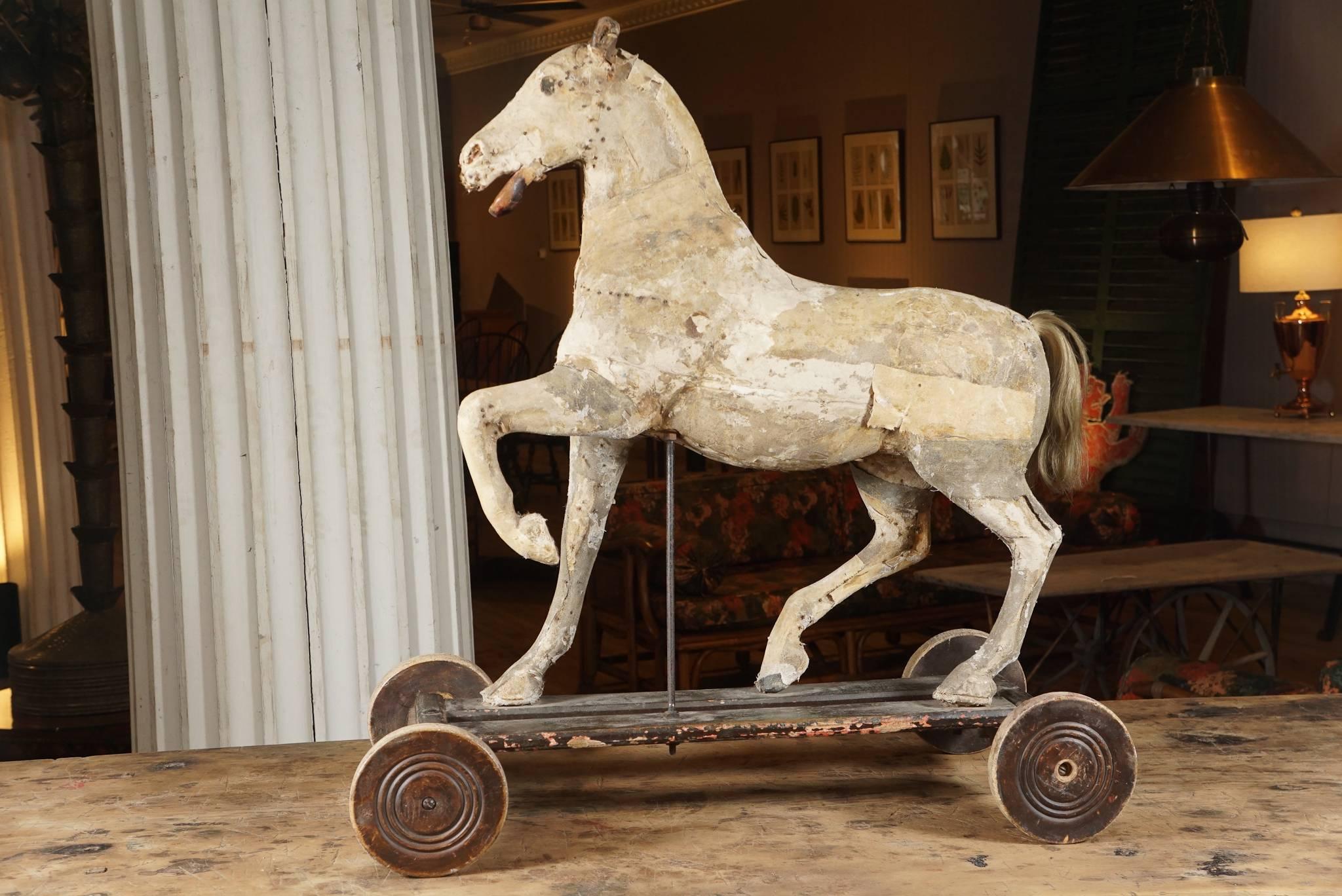 18th century sculpture of a prancing horse, exquisite form, ancient velum colored surface of hide and nail construction, wood carved hooves and jaw, leather ear, white horsehair tail, on wood wheeled base in original brown painted worn surface.