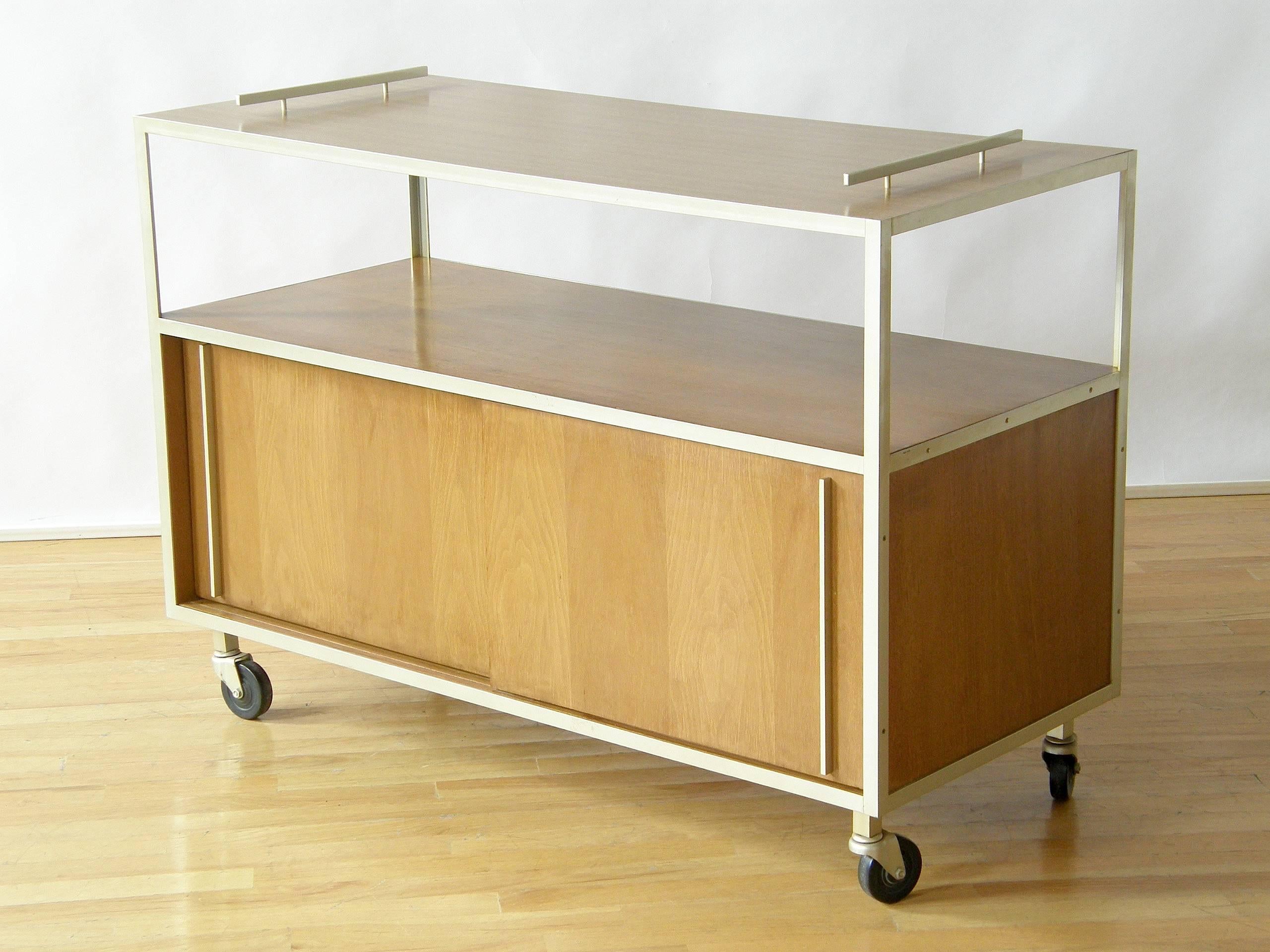 Utilitarian, late 1950s serving cart with a laminated top surface for protection against spillage, an open shelf below that and a large, closed storage area in the bottom section with sliding doors.

Please contact us if you have any questions.