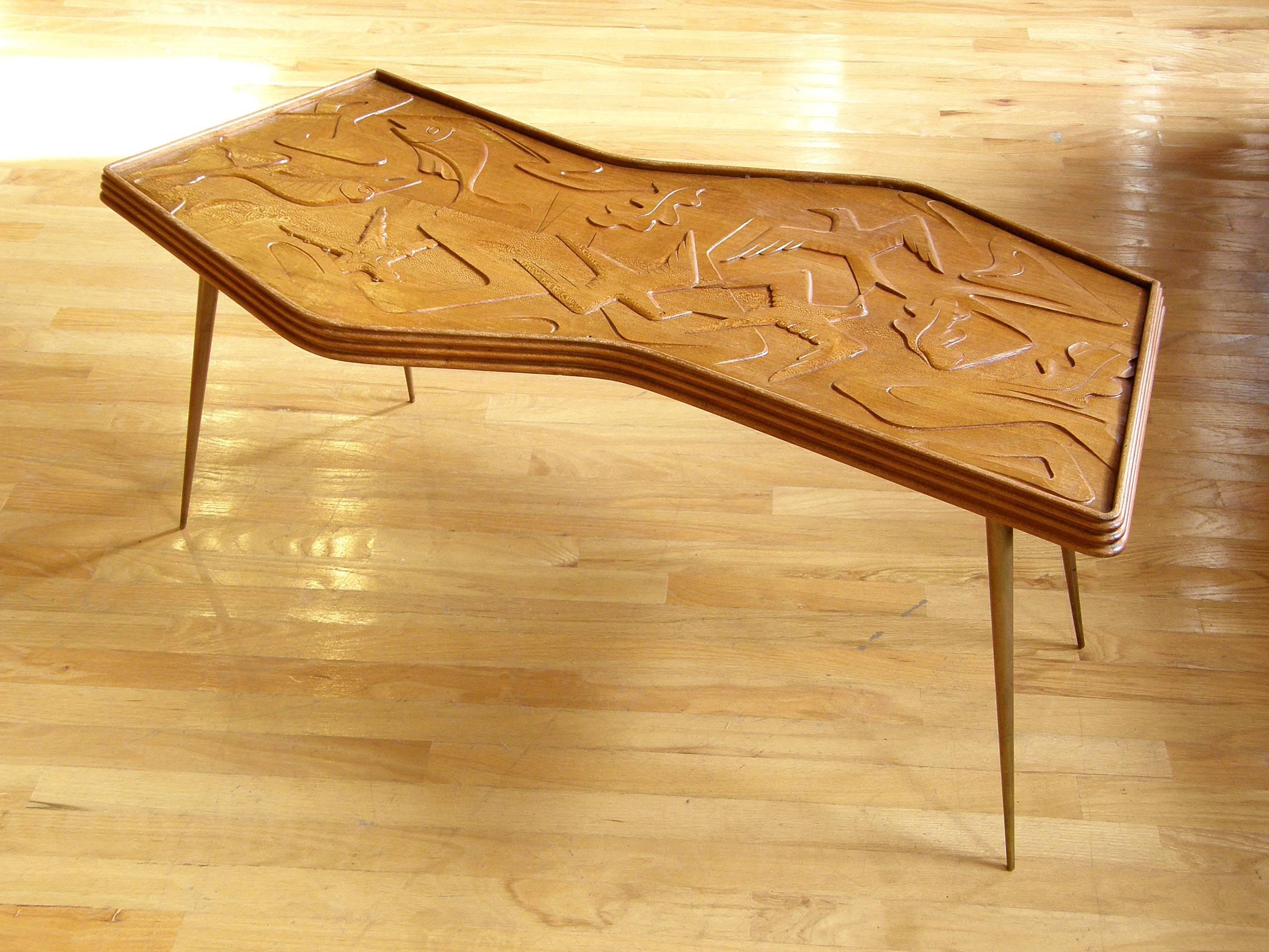 Sculptural zigzag coffee table in mahogany with slender, tapered brass legs. It has a stepped rim around the edges with rounded corners. The top is a wonderfully carved relief sculpture with stylized, modernist birds, leaves, fish and suggestions of