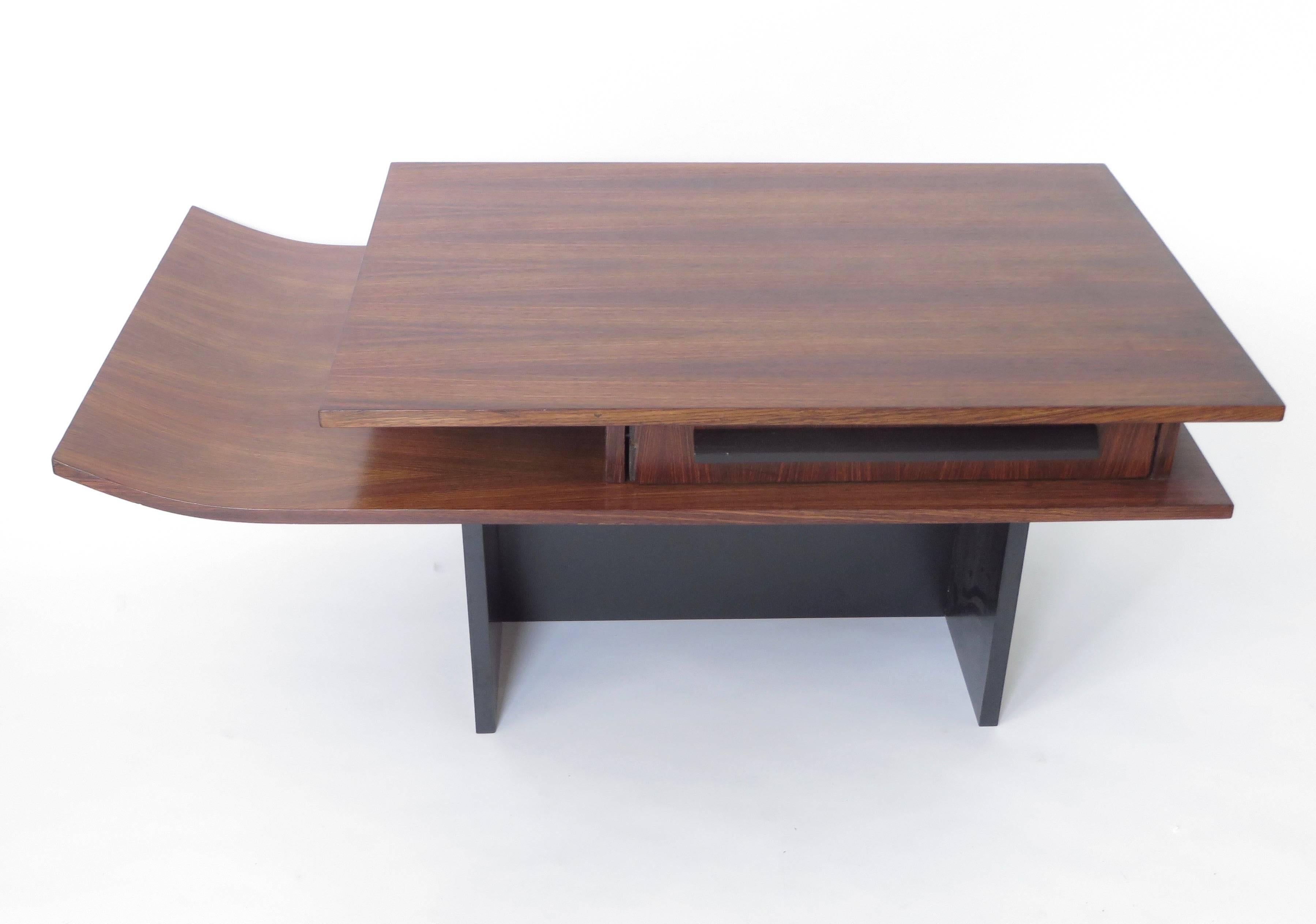 An coffee table of unknown designer made of two levels of rosewood, one level with a gently sloping upward curve. One drawer with ebonized rosewood drawer pull sitting on black laminate base. 
A unique feature of the drawer is that it slides through