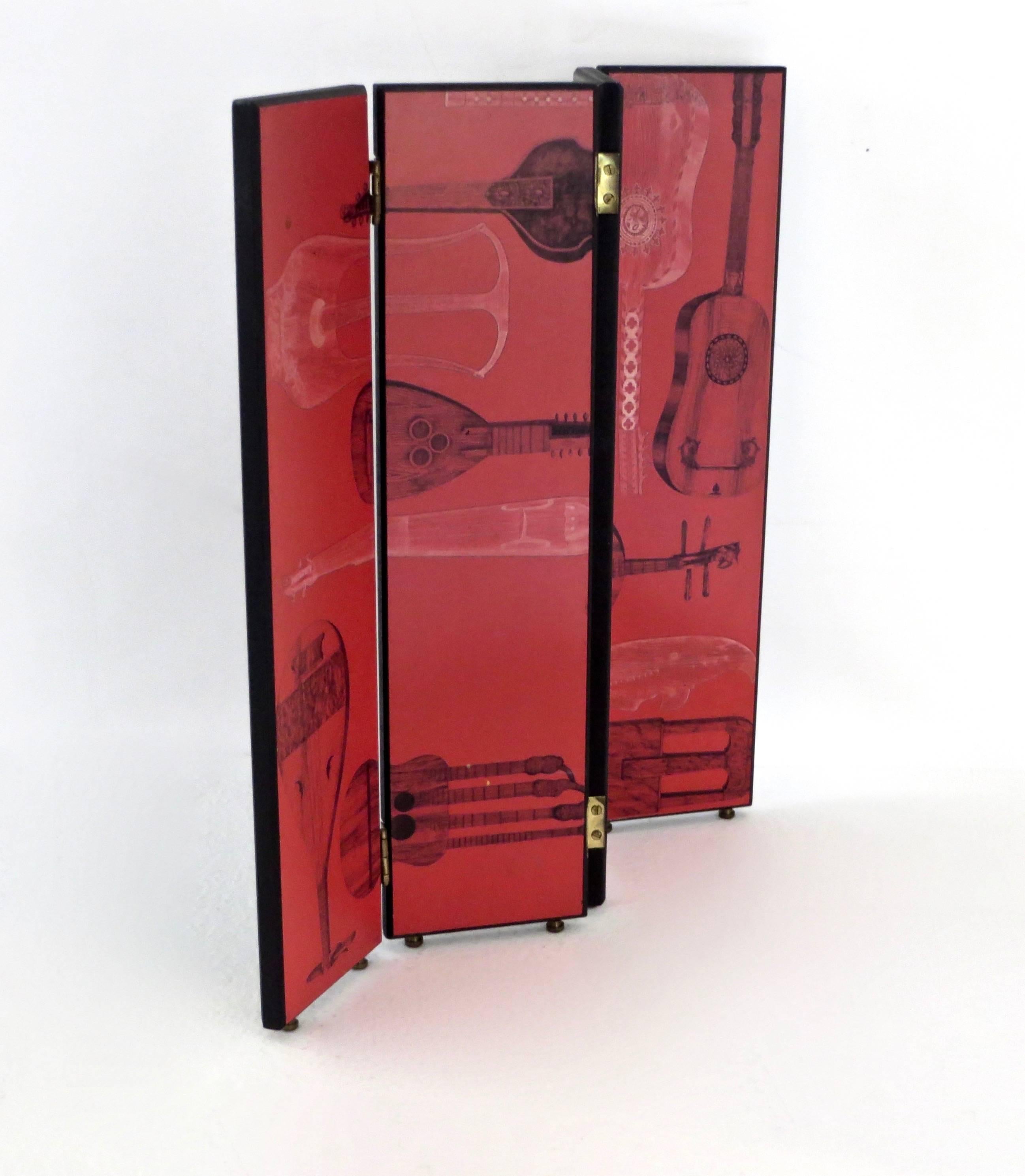 A miniature folding screen by Piero Fornasetti with musical instrument motifs on the front in red and black and the reverse side showing black background with white sun motif.
Miniature brass feet in full example of the large-scale