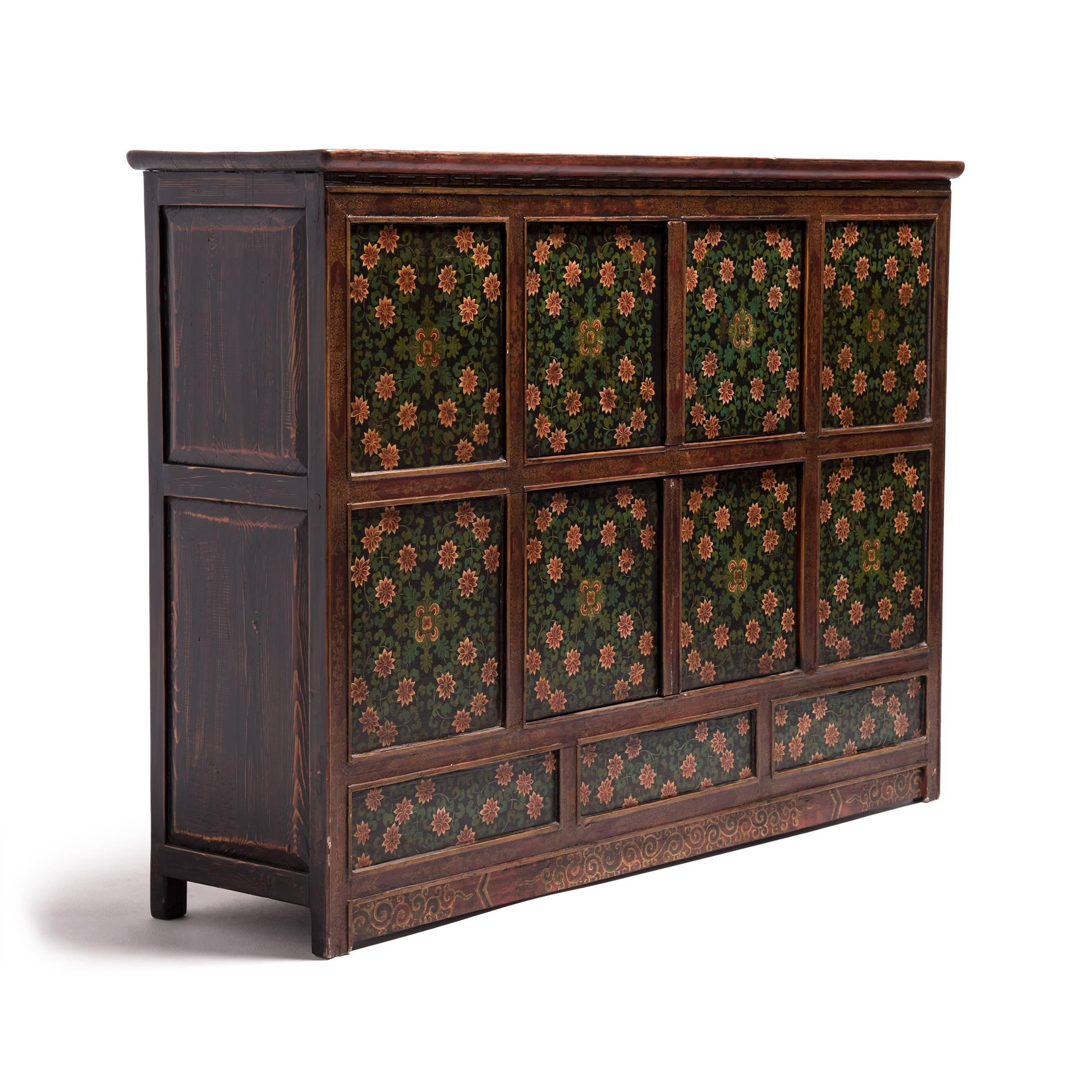 Influenced by early 19th century Tibetan painted wood furniture and panels created in monasteries, this early 20th century cabinet is lavishly decorated with painted floral motifs that reference Buddhist iconography. Paneled doors depict trailing