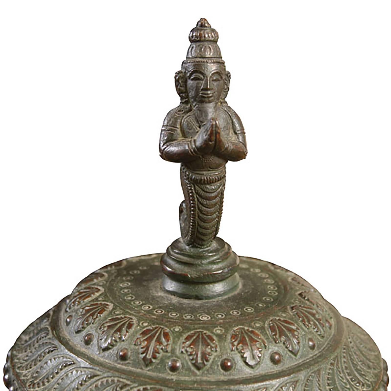 This bronze vessel was cast in India over a century ago. The artisan’s talent and craftsmanship are impressive and evident in each beautiful detail. The lid is ringed by tiny leaves and crowned with a figure whose hands are raised in prayer. The
