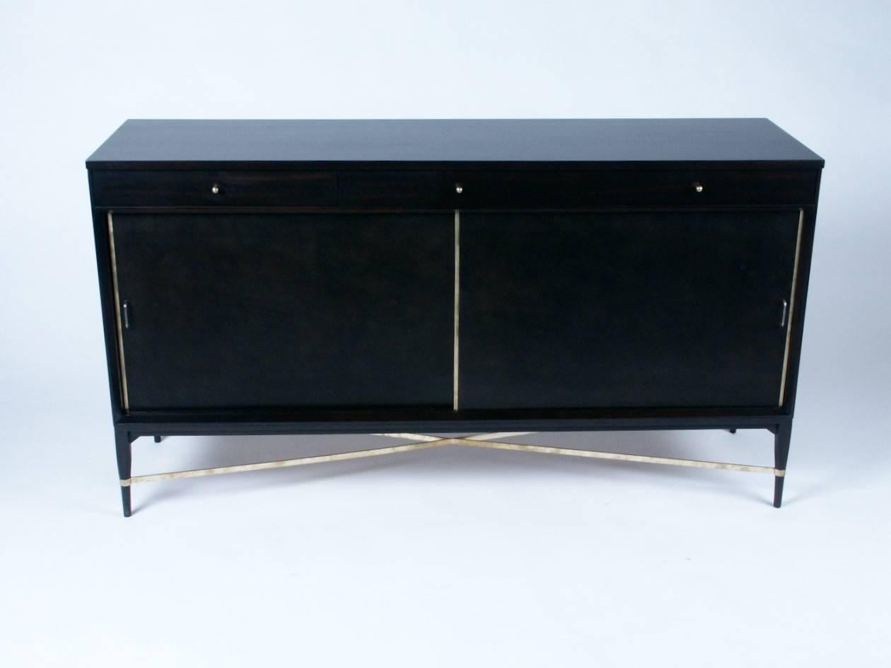Paul McCobb sideboard, three drawers at top, sliding leather doors with brass trim, and adjustable shelves, brass X stretcher between tapered legs. Currently being refinished in dark espresso.