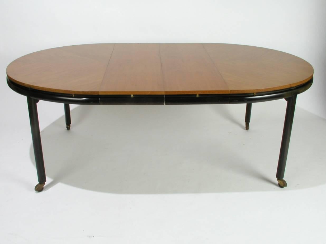 Oval dining table designed by Winsor White and Michael Taylor for Baker's New World collection, circa 1950s, ebonized finish on legs, brass castors, top is given floating effect by vertical brass cylinders set into frame, top is medium light wood