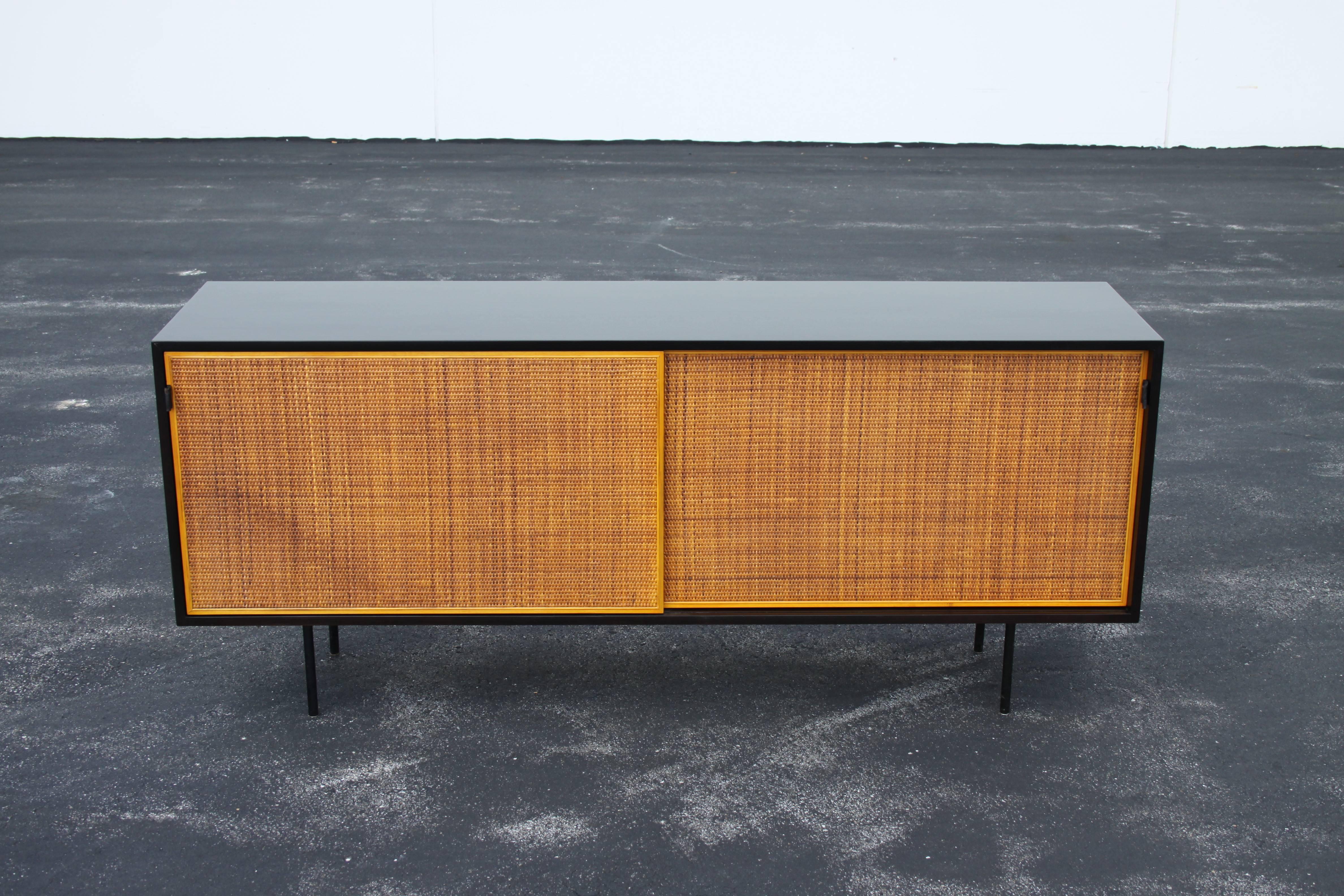 Early Florence Knoll walnut sideboard or credenza with black ebony stain, two sliding doors with leather tabs, model number 116, Interior lacquered off-white with seven adjustable oak shelves, black iron tubular legs. Credenza to be refinished,