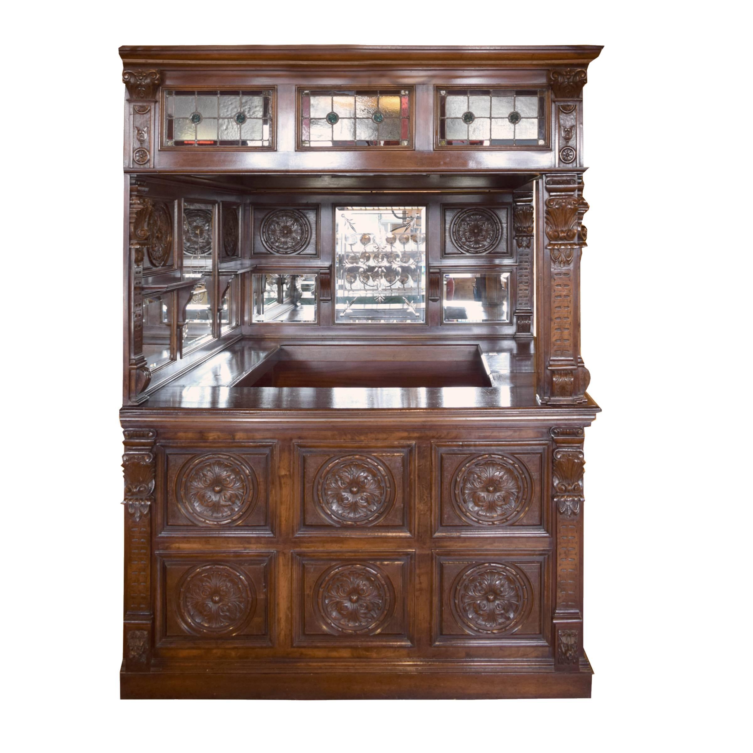 A highly decorated English wood bar with carved floral panels, beveled and cut-glass mirrors, stained glass panels along the top, and a stained glass skylight depicting a bird. The interior features two low shelves that extend along two sides, three
