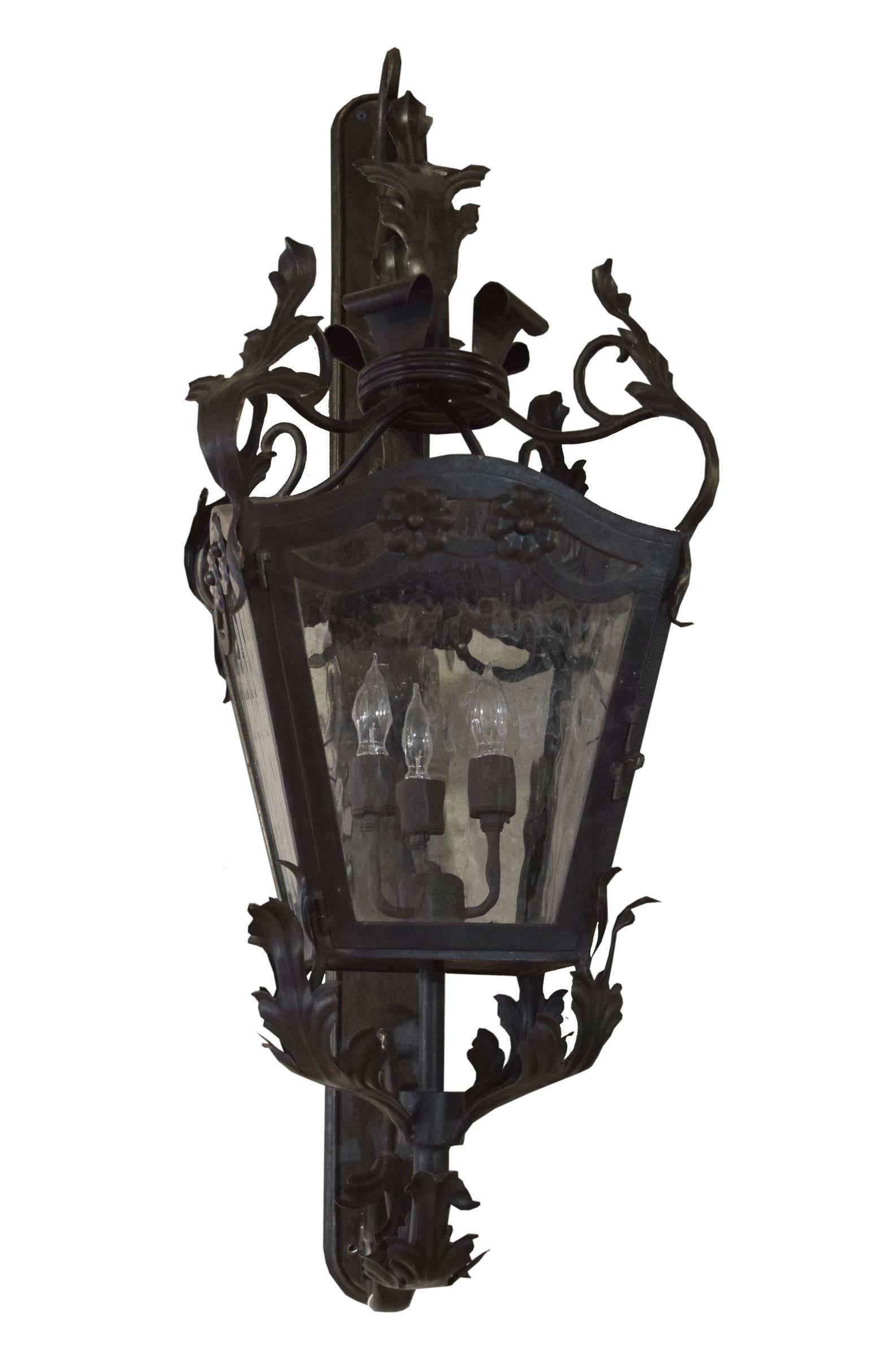 A large wrought iron wall mounted lantern from Argentina with four waterfall glass sides and three lights. Adorned with small flowers and a crown, scrolling vines and leaves support the fixture from above and below.