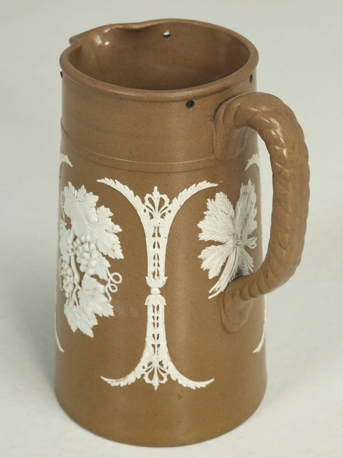 English stoneware pitcher in a most unusual brown color and decorated with white berries, grapes and sheaves of wheat, all in a relief. The antique English stoneware pitcher was made in 1848. There are four tiny holes at the very top and we are