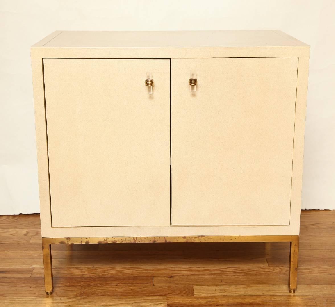 A pair of side tables, the top sides and cabinet doors finished in white lacquered linen. The cabinet doors with brass and lucite pulls concealing interior with single shelf, supported by open square bronze frame bases. Circa 1975. 
