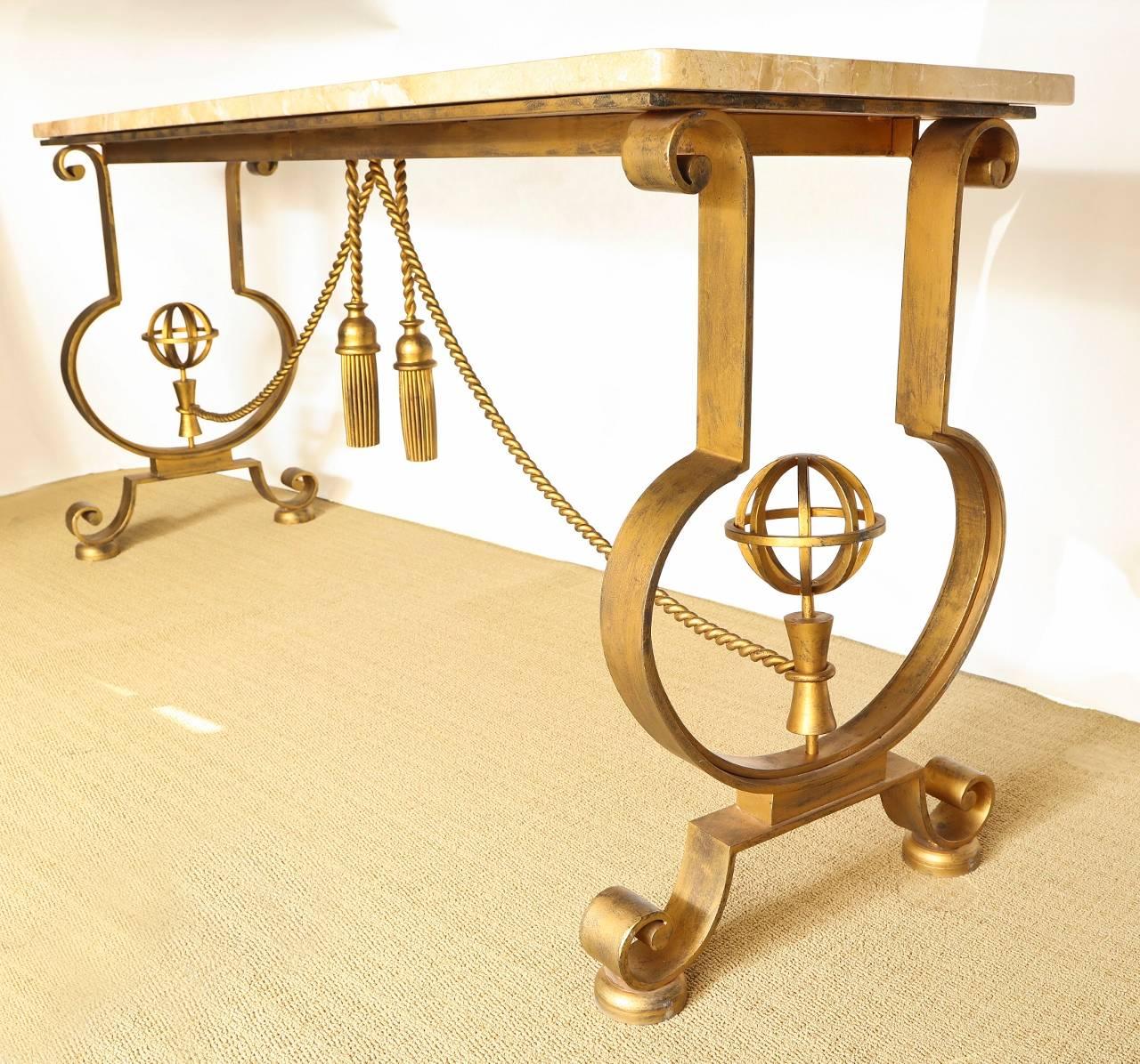 A French forged iron gilt console in the manner of Gilbert Poillerat. The marble-top table has a support at either end with open work design featuring armolary sphere above splayed legs, the ends joined by stretcher in the form of draped rope with