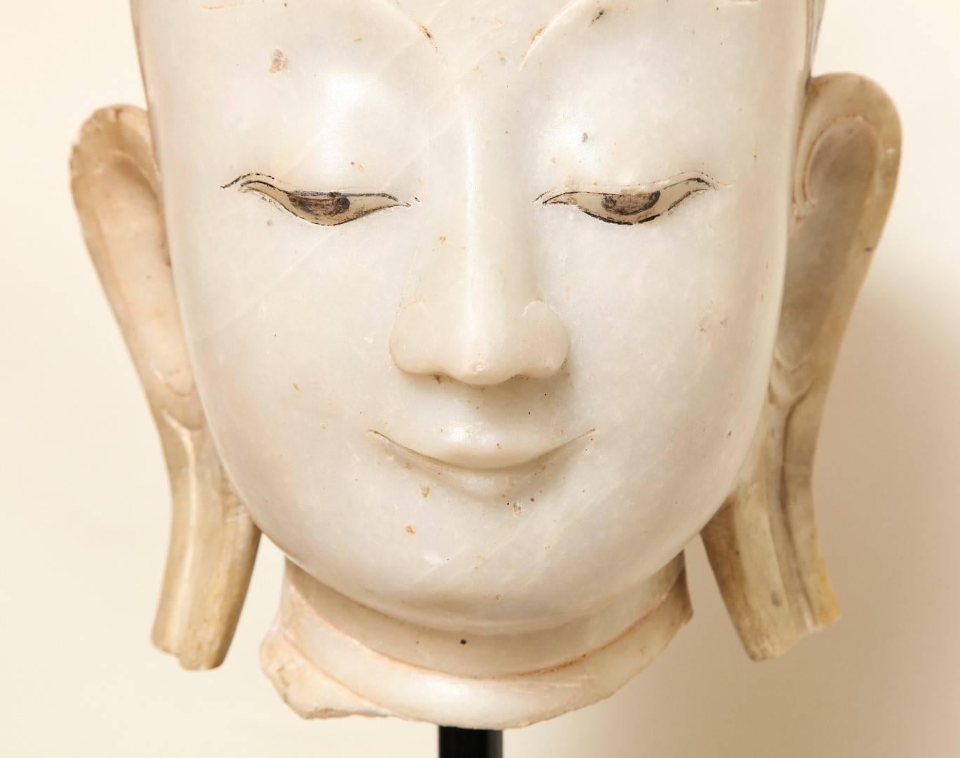 The life-sized marble Buddha head sympathetically carved, the mouth bearing a hint of a smile. The hair and facial features showing traces of gilding and paint. Museum mounted, Burmese.