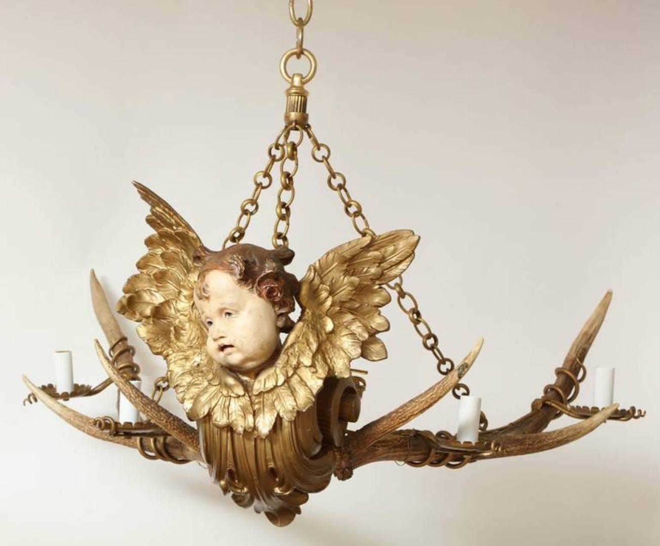 An Austrian four-light chandelier with a rack of natural deer antlers mounted with a carved wood figurative element of a winged cherub with fish scale body/tail. The natural antlers have iron tendrils encircling it and metal candle cups, now
