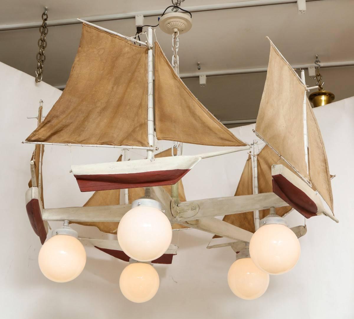 A sailboat form chandelier, the off-white painted wooden frame in the form of a propeller issuing five arms with affixed sailboat with linen mast, the underside of each arm suspending a globe form light, Edison socket, 40 watt maximum recommended 
