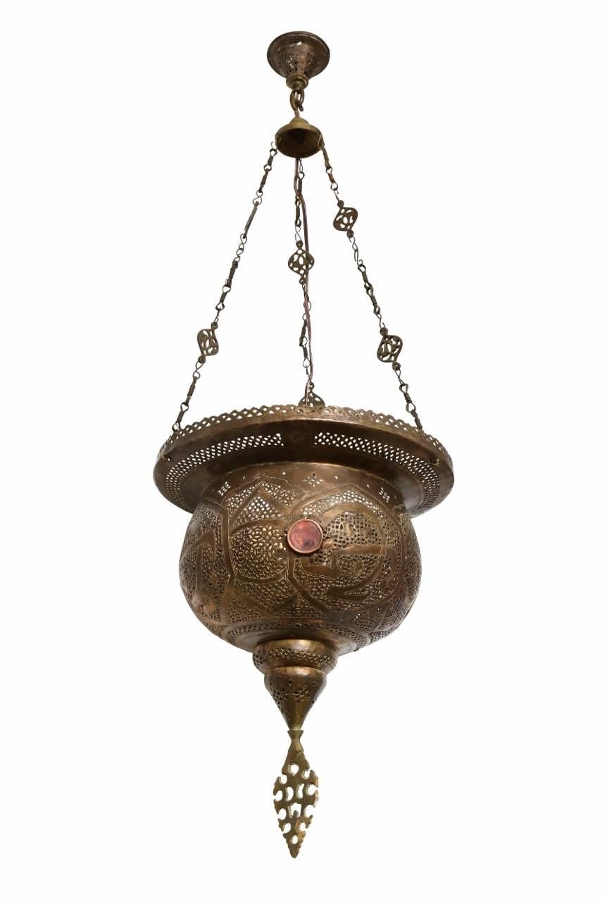A vase shaped Morrocan pierced brass ceiling fixture with round colored glass elements in body suspended from three lengths from brass chain. The interior fitted with electric socket