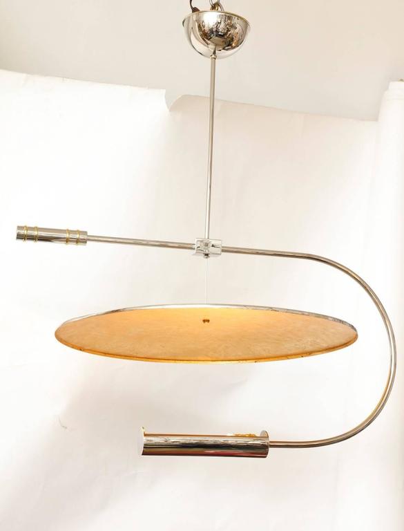 A silver-nickel contemporary design Bauhaus inspired ceiling light with curved arm issuing light from open cylindrical element onto shallow dish with 24K gold leaf on reflective surface, the dish suspended from wire. Asymmetrical design features