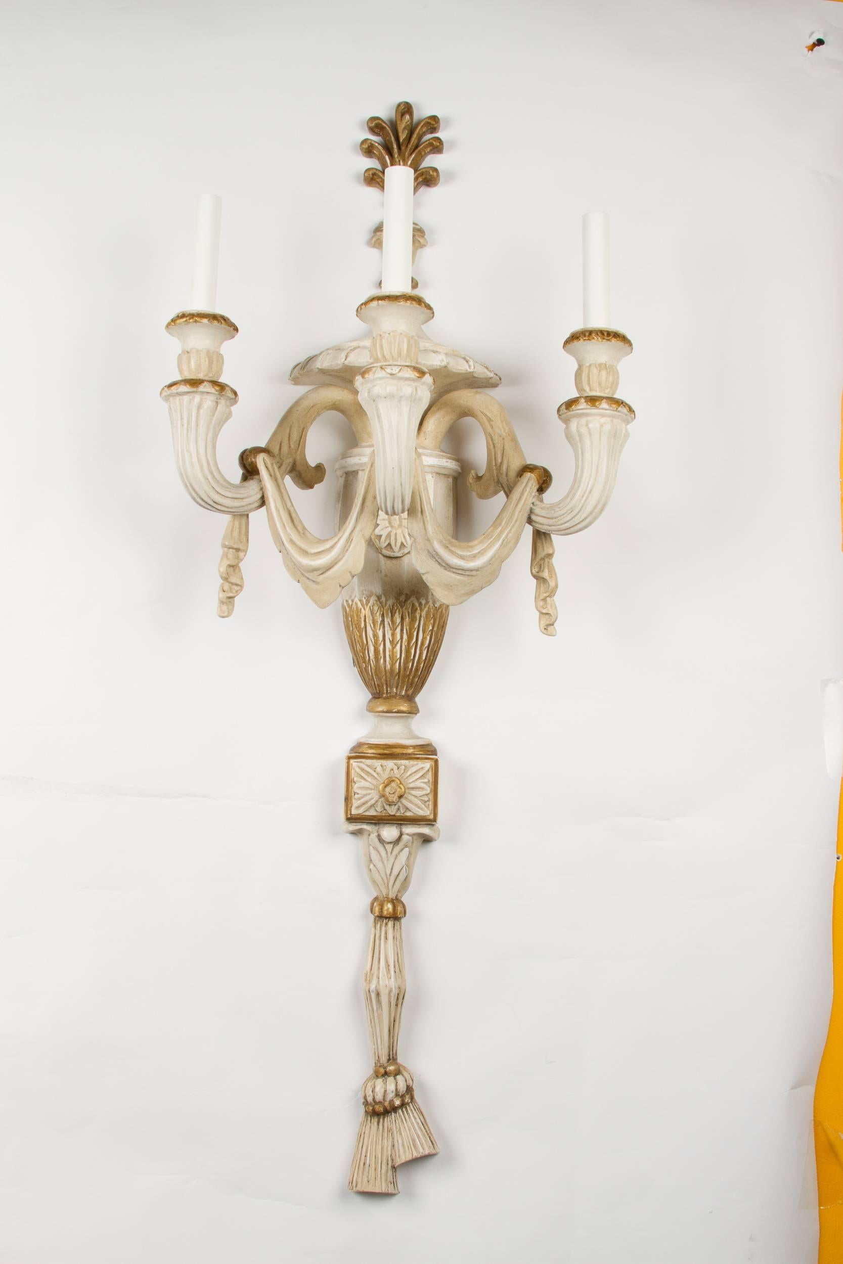 A pair of cream painted and parcel-gilt wall appliques, 3 light neoclassical design with vase shaped back plate having acanthus leaf and paterae decoration and issuing three scrolled candle-arms suspending trompe l'oeil drapery, terminating in