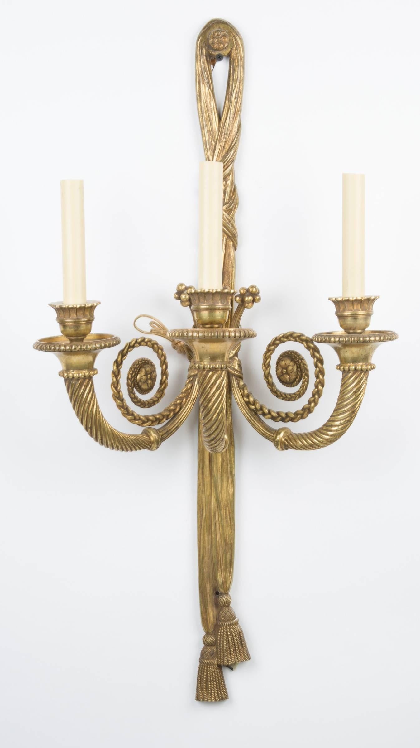 A pair of French Louis XVI-style three-light sconces, the backs in the form of draped cord with cross bands terminating in tassels, each candle arm with spiral twist fluting.