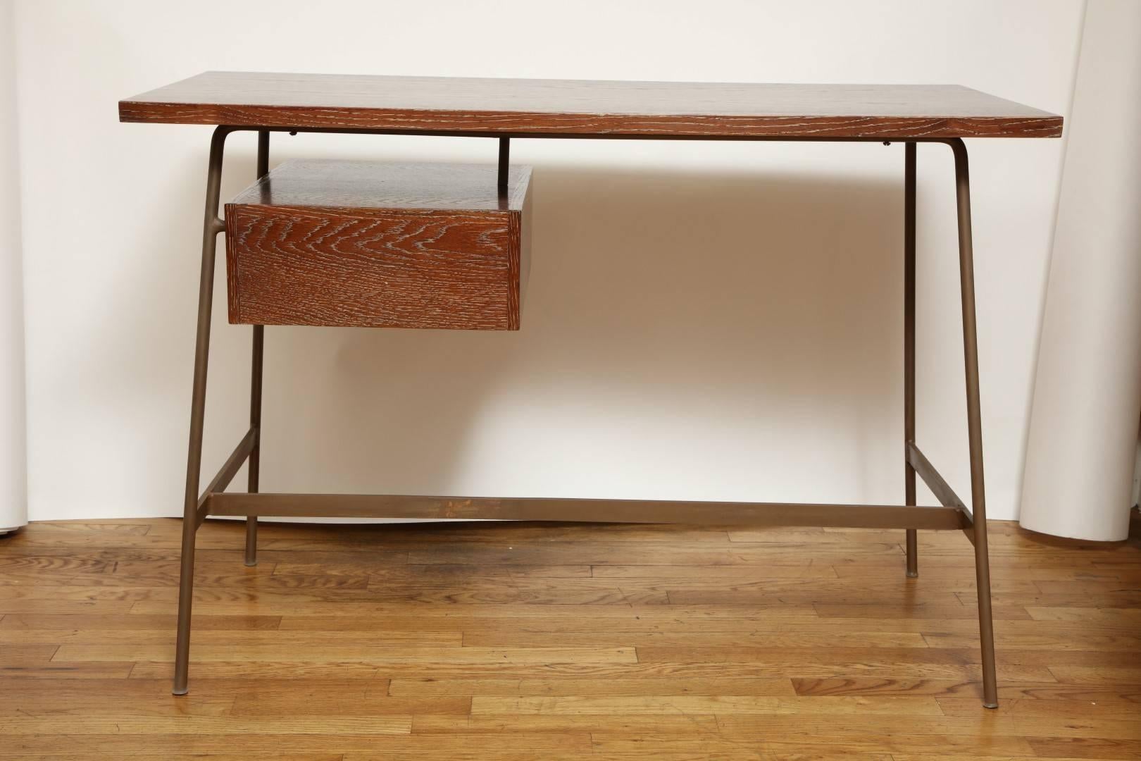 A new midcentury style cerused oak desk with floating drawer at right of the underside with original leather tab pull and supported by four splayed iron legs joined by H-shaped stretcher.