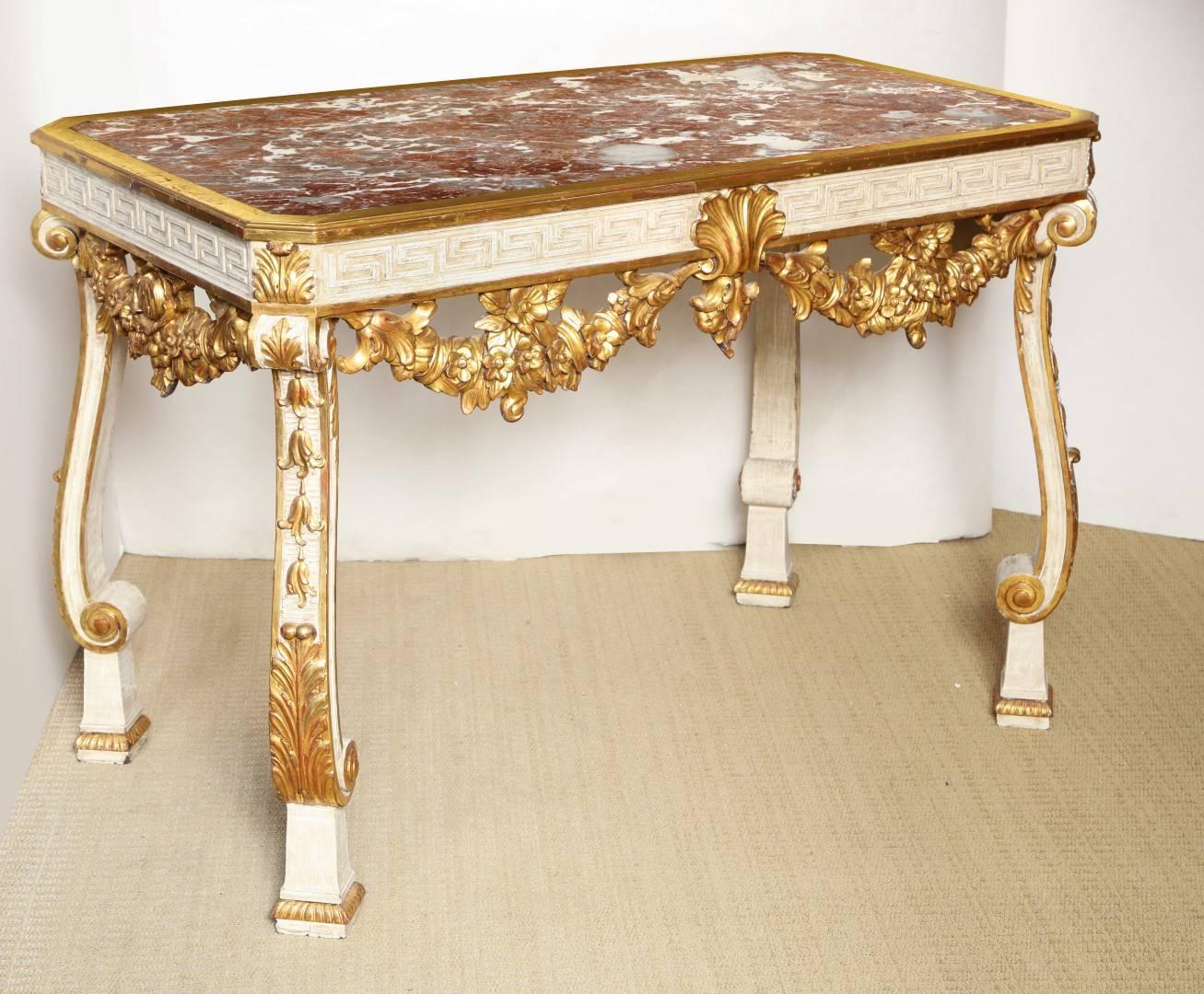 A George II style parcel-gilt and cream painted console table, the Rouge Des Pyrenees marble top inset into gilt frame with canted corners, resting on a Greek key carved freize centering a gilded scallop shell issuing lush festoons, the foliate legs