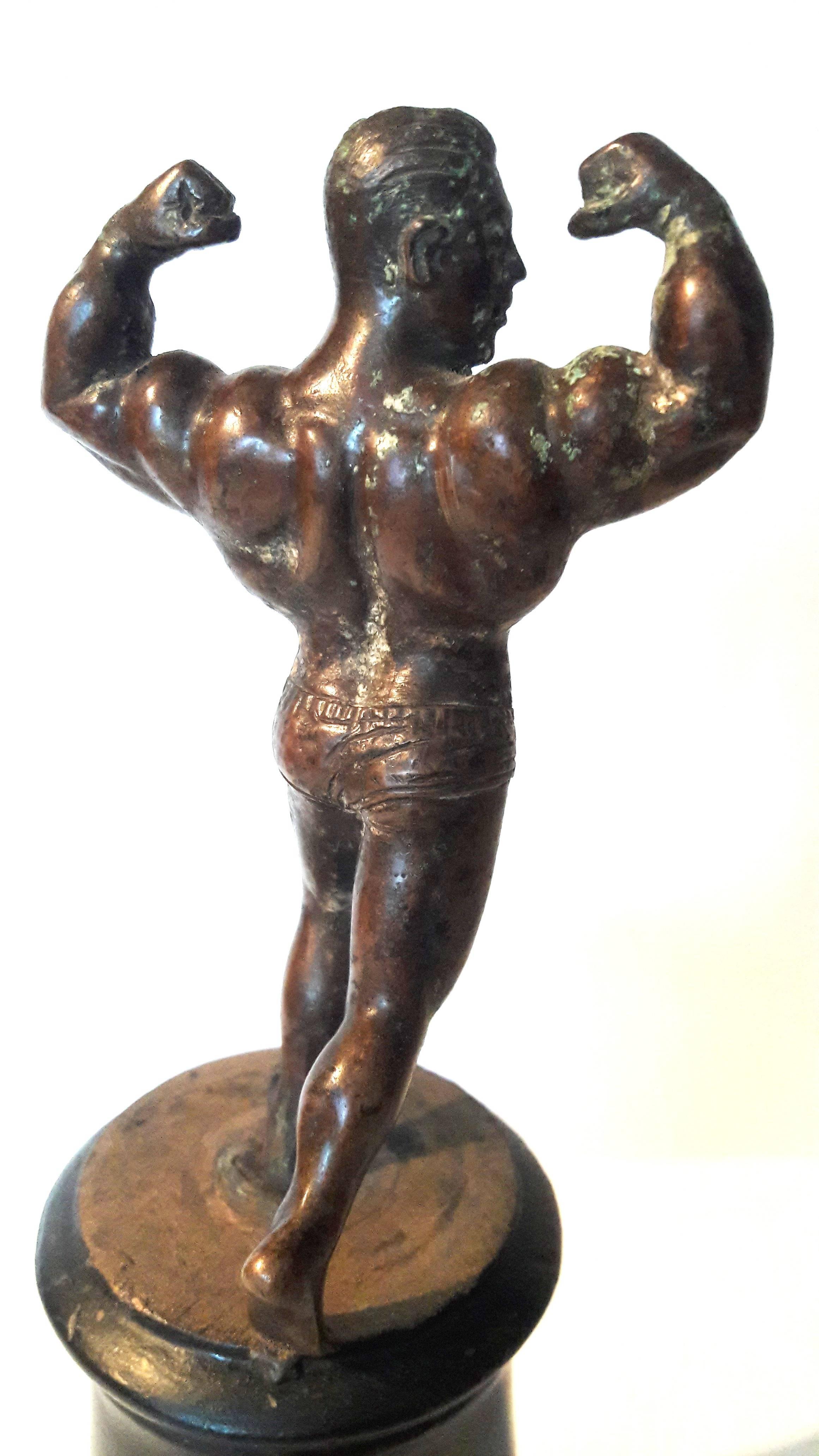 Une petite statue d'homme musclé en bronze sur un socle en bois noir.
Mesures : base de 9 1/2