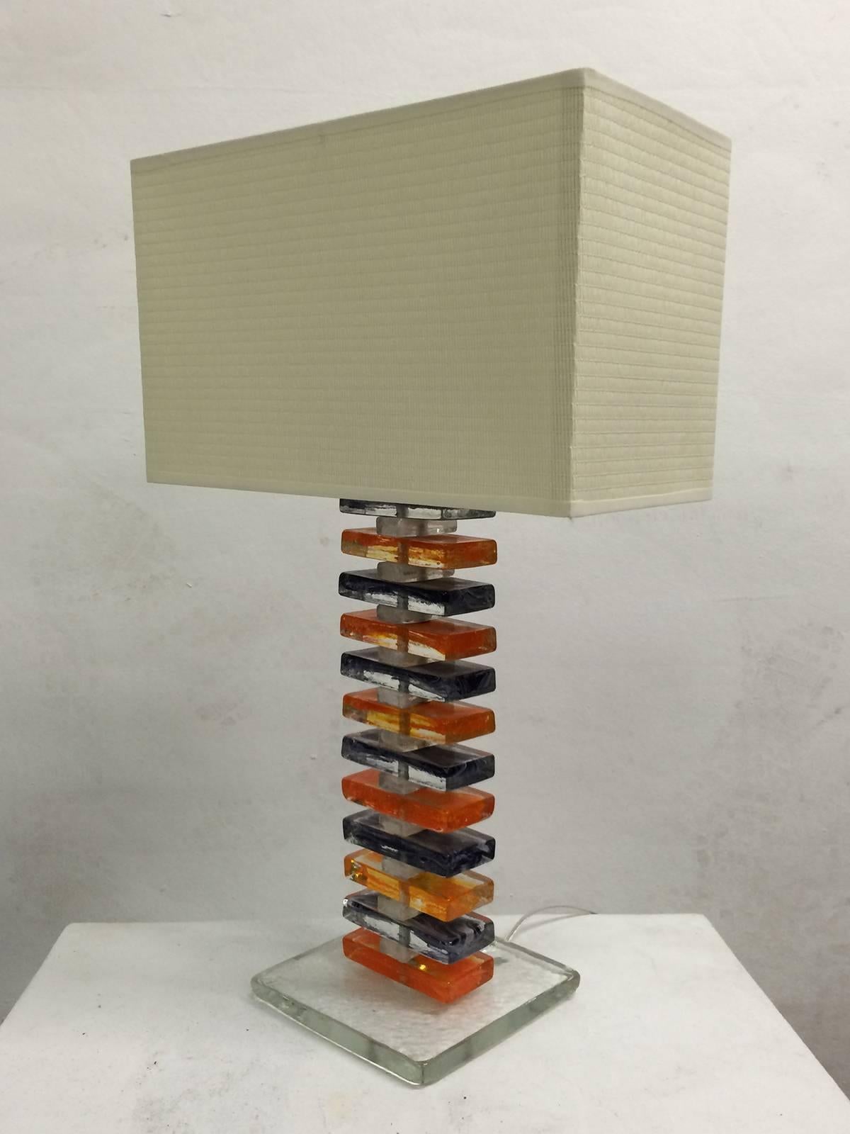 A pair of table lamps in stacked colored glass alternating clear, orange and purple. The base alternates small and large pieces almost like a spine. Finished with a rectangular shade, the lamp has a very geometric feel.  Shade is 16 wide by 8 deep